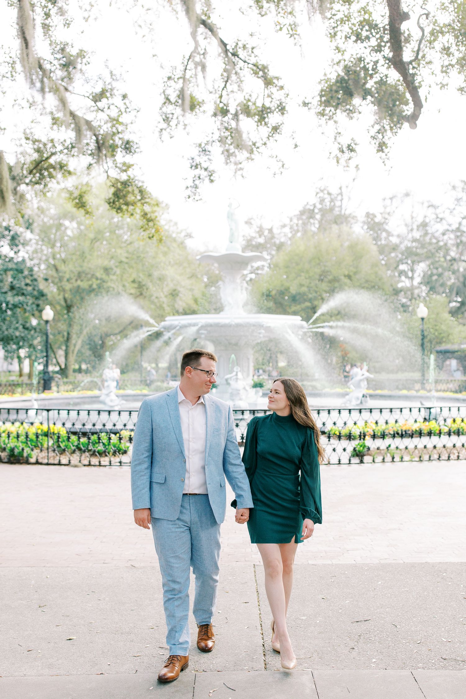 A couple poses together near the beautiful Forsyth Park fountain in a park setting with moss-covered trees.