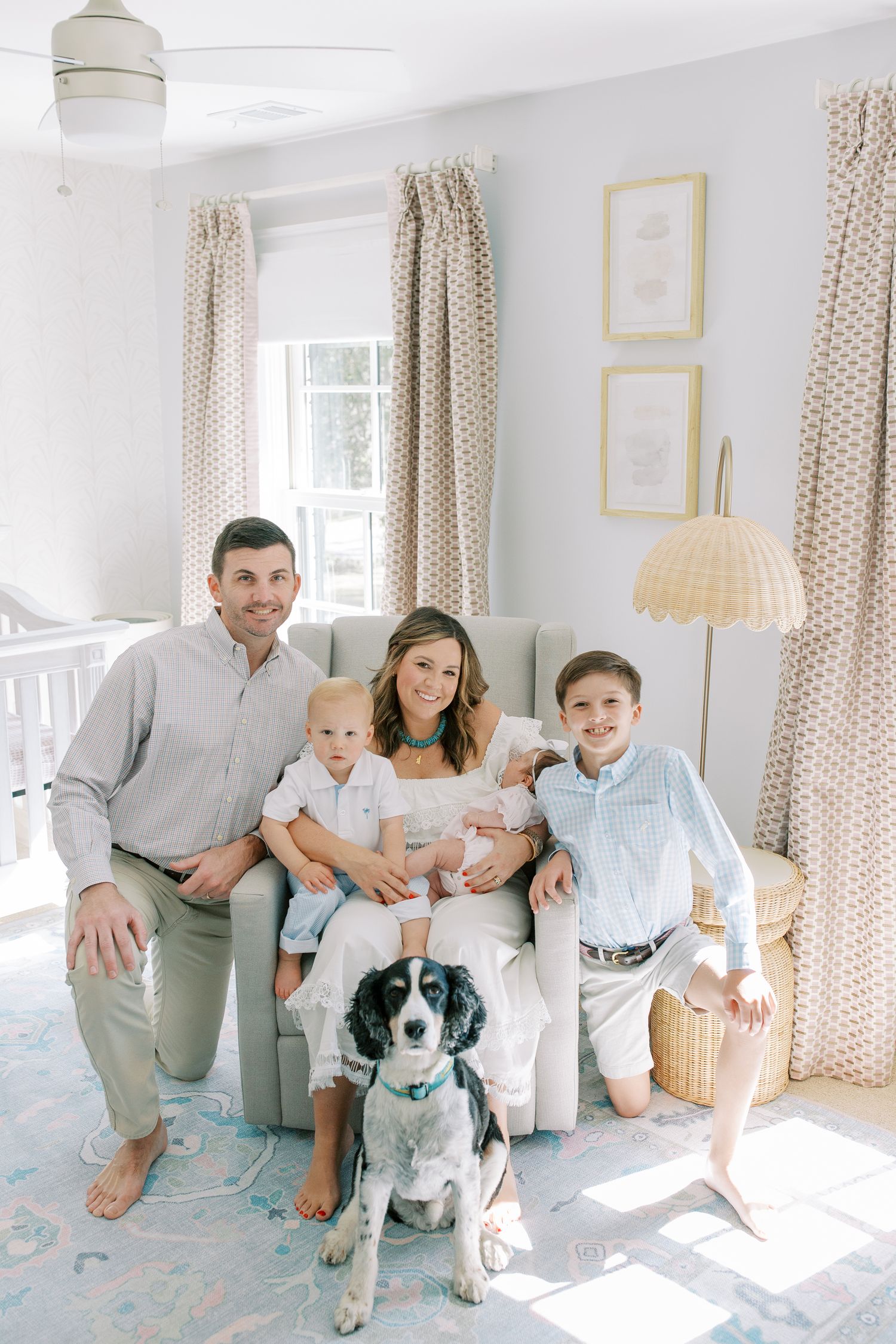 A bright sunlit room with polka dot curtains features a family gathered on a chair with their black and white dog.