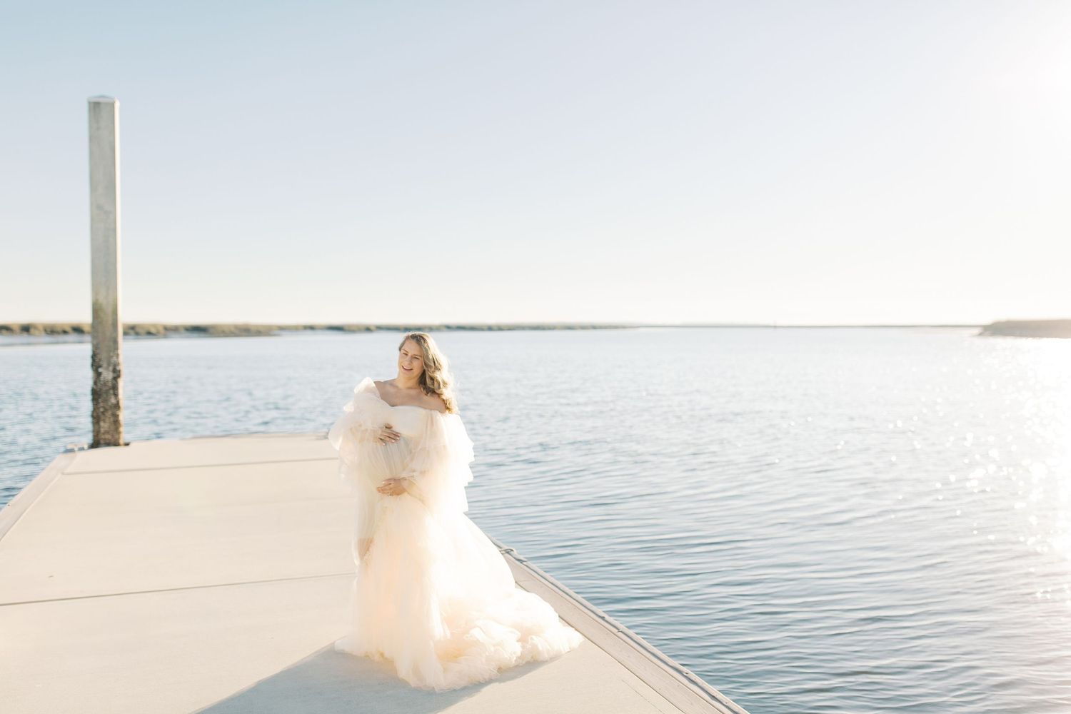 A couple in wedding attire embraces on a wooden dock by a serene lake on a bright sunny day.