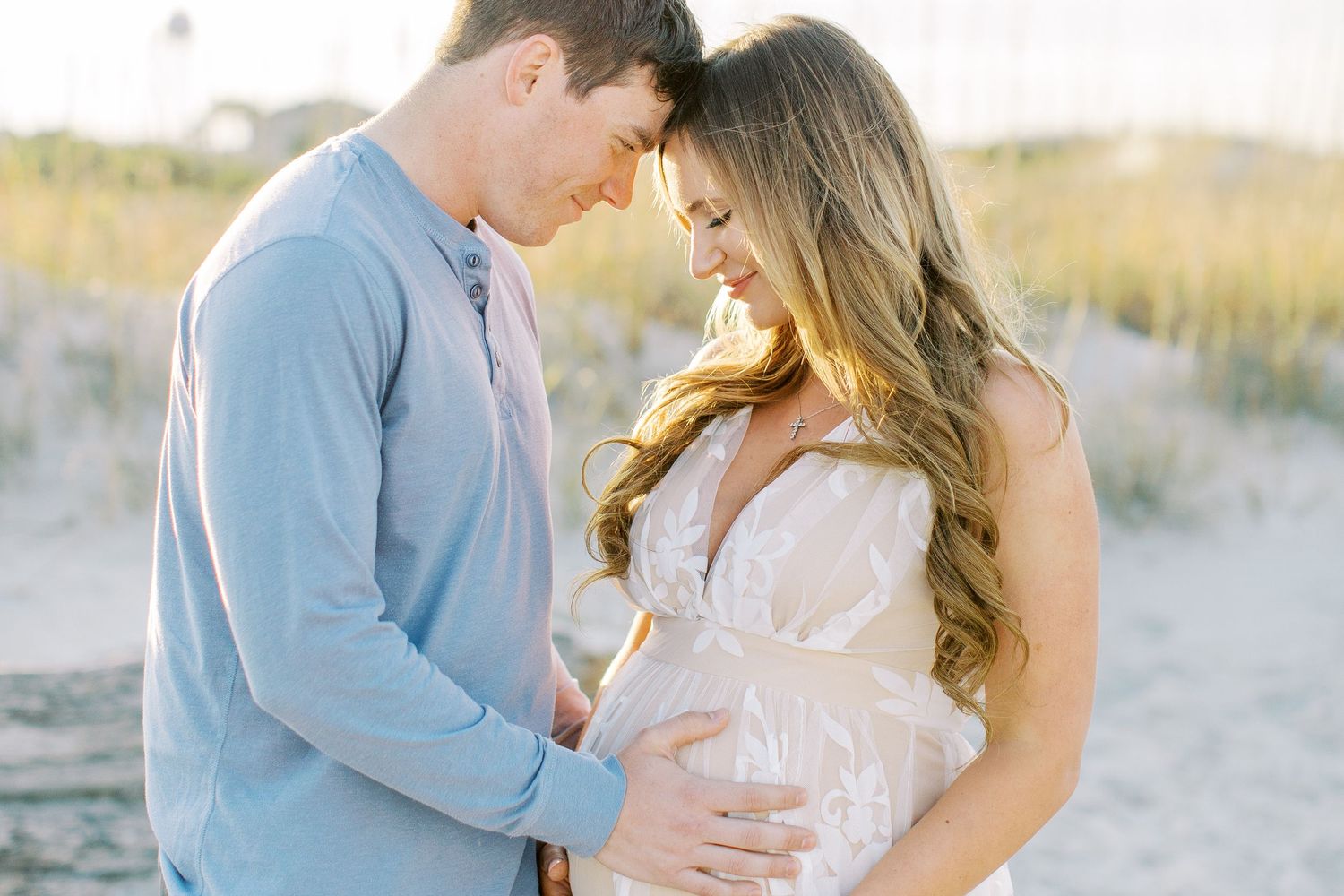 A couple shares an intimate moment during maternity session on tybee beach with her wearing a white dress and him in a light blue sweater.