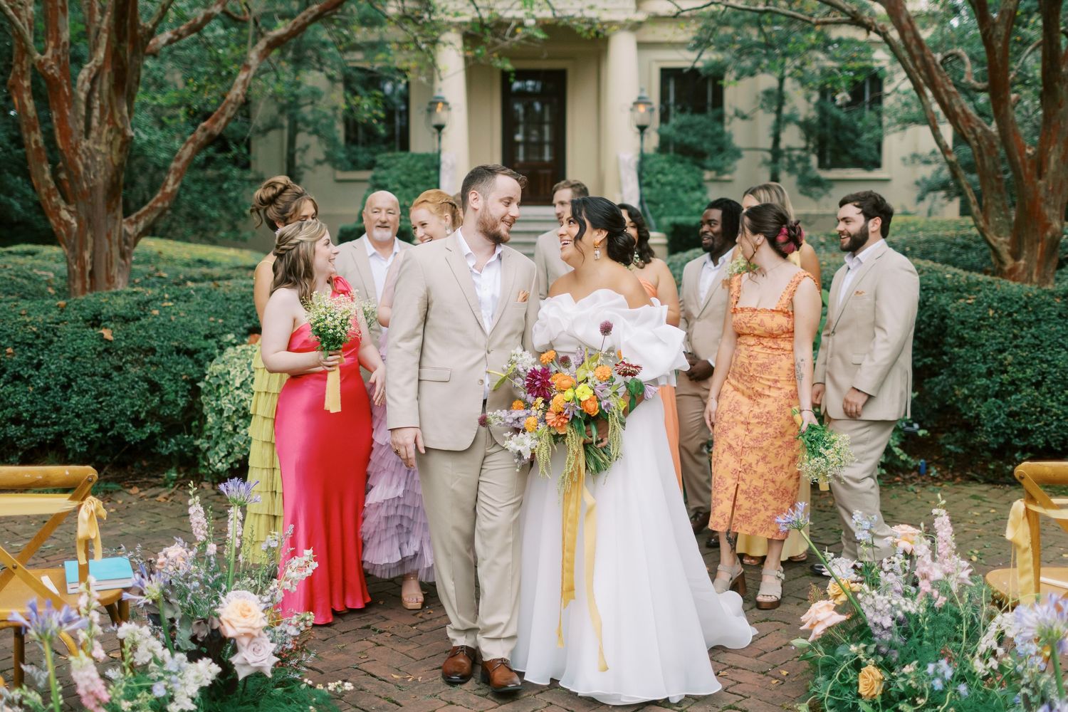 Wedding party gathered outdoors wearing pastel and coral tones amidst garden greenery.