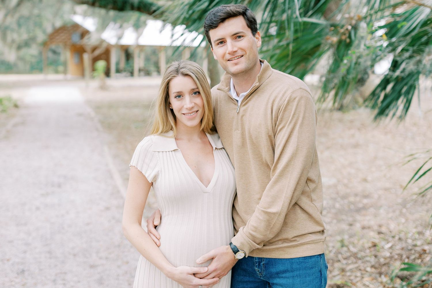 A couple in light colored clothing embraces outdoors among palm trees and foliage during a photo session.