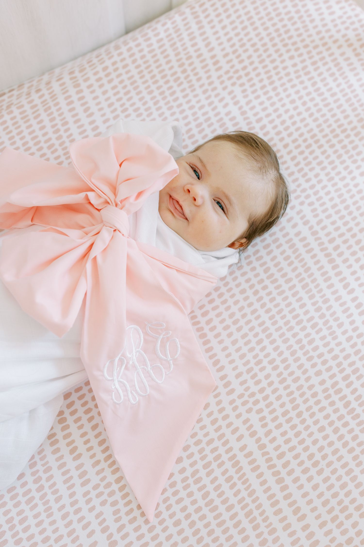 A baby swaddled in white lies on patterned fabric with a large monogrammed pink bow.