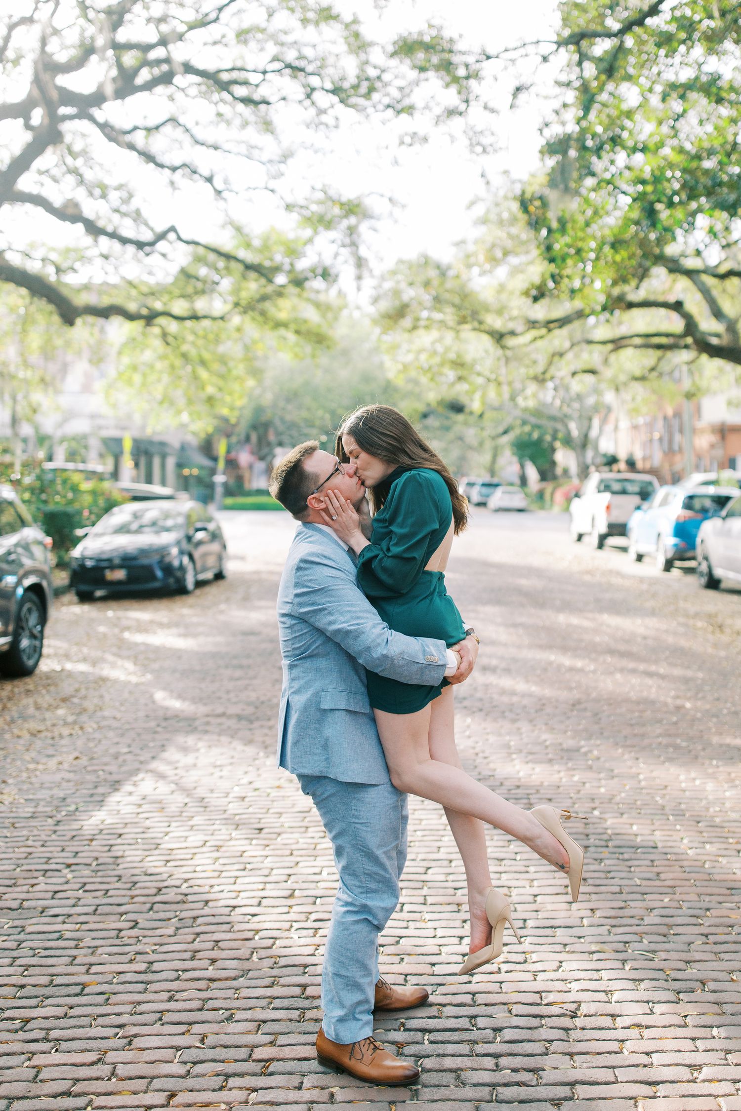 A romantic moment captured on a cobblestone street with overhanging trees in the background.