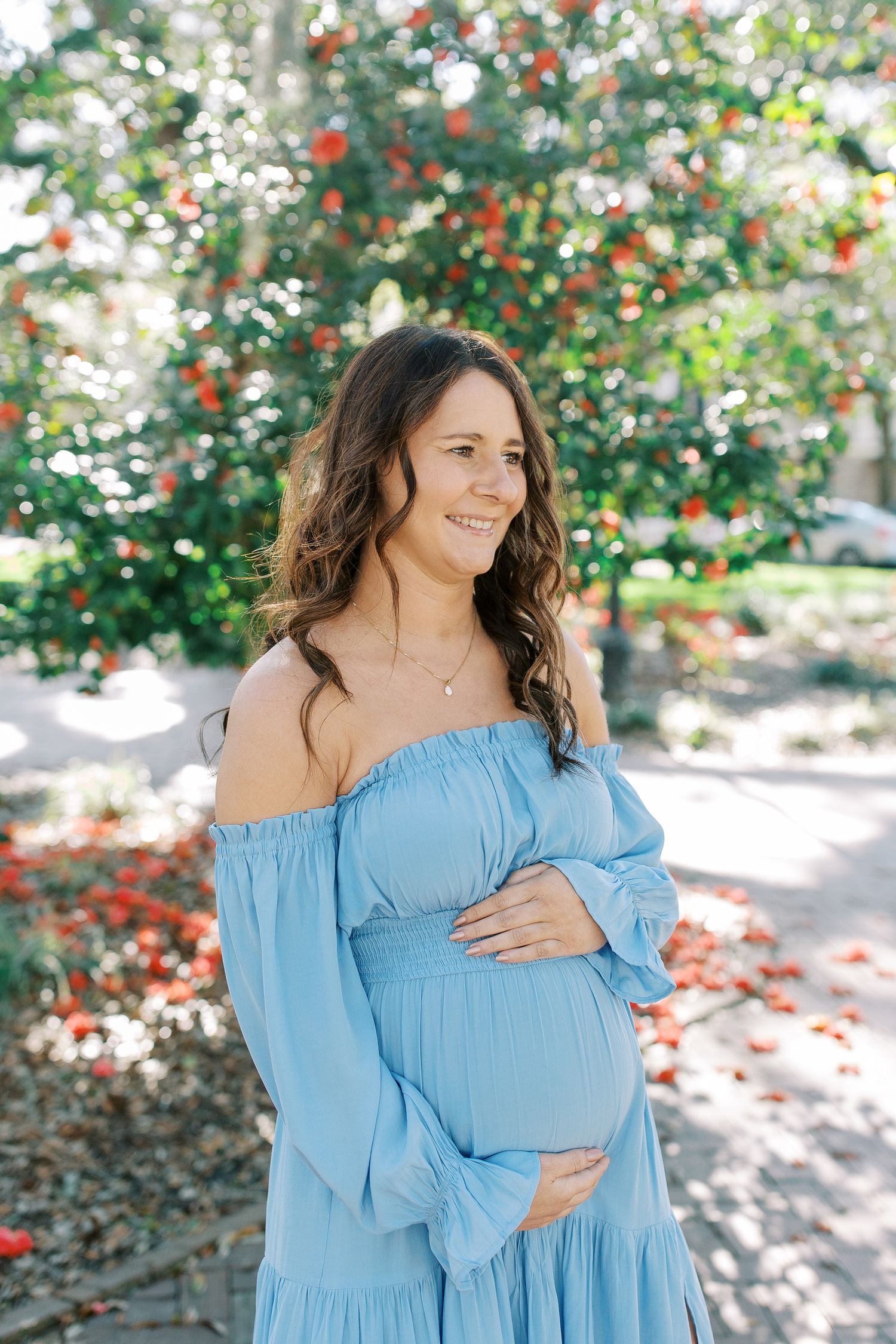 Expectant woman in light blue off-shoulder maxi dress poses outdoors with red berries and greenery in background.