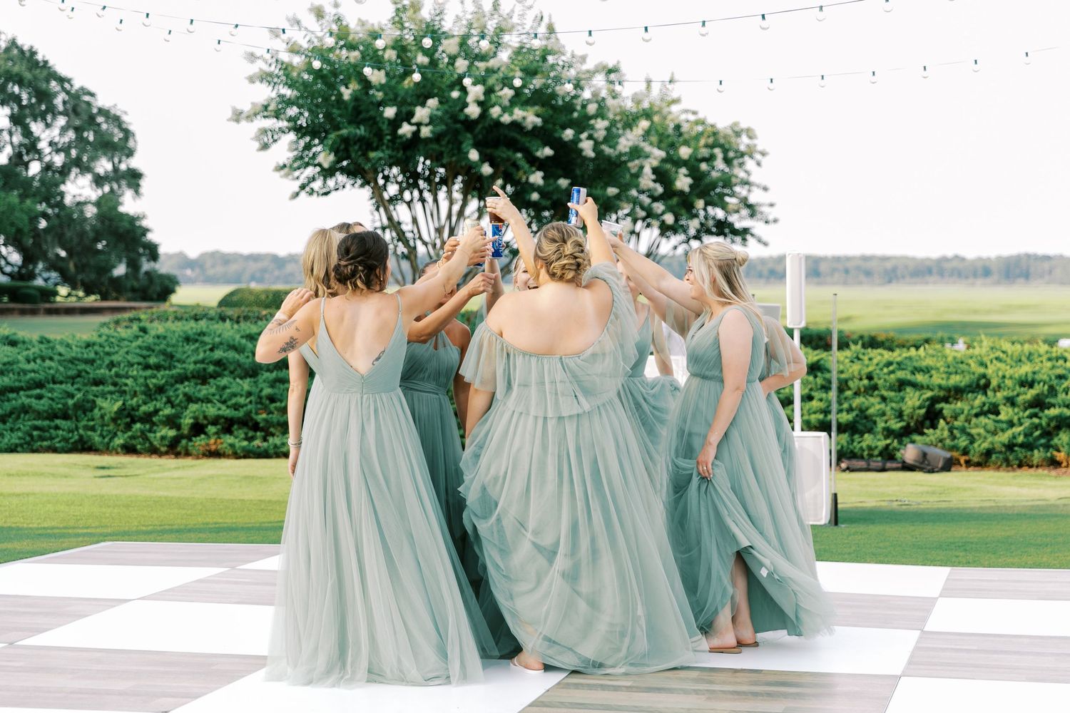 People in flowing sage green bridesmaid dresses dance together on an outdoor dance floor during a wedding reception.