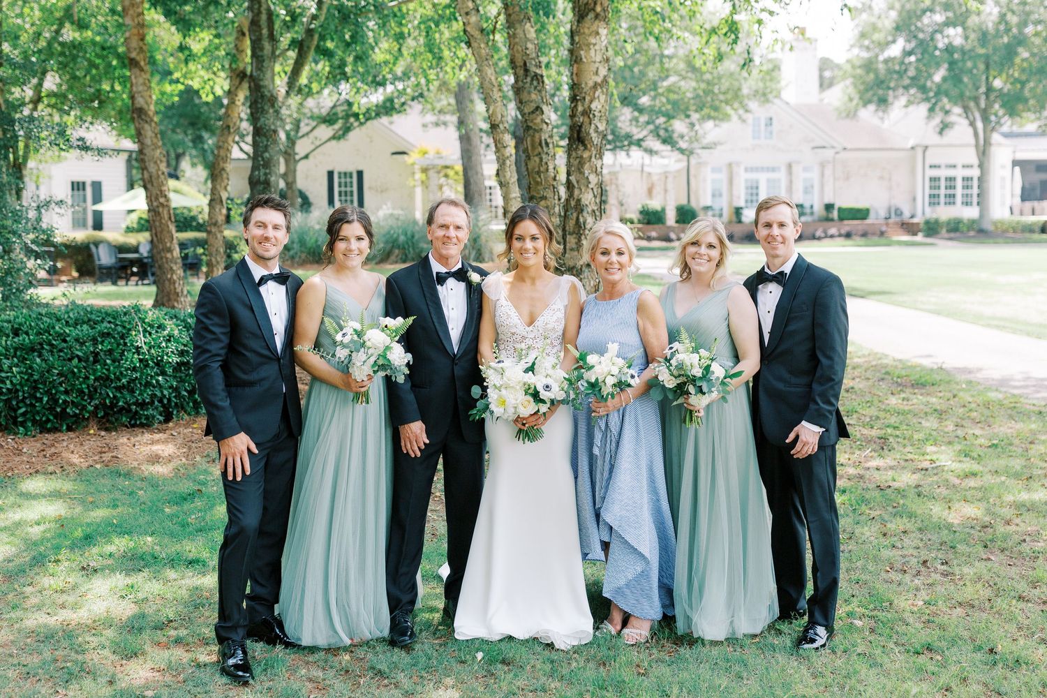Wedding party posed together outdoors wearing blue dresses and black tuxedos.