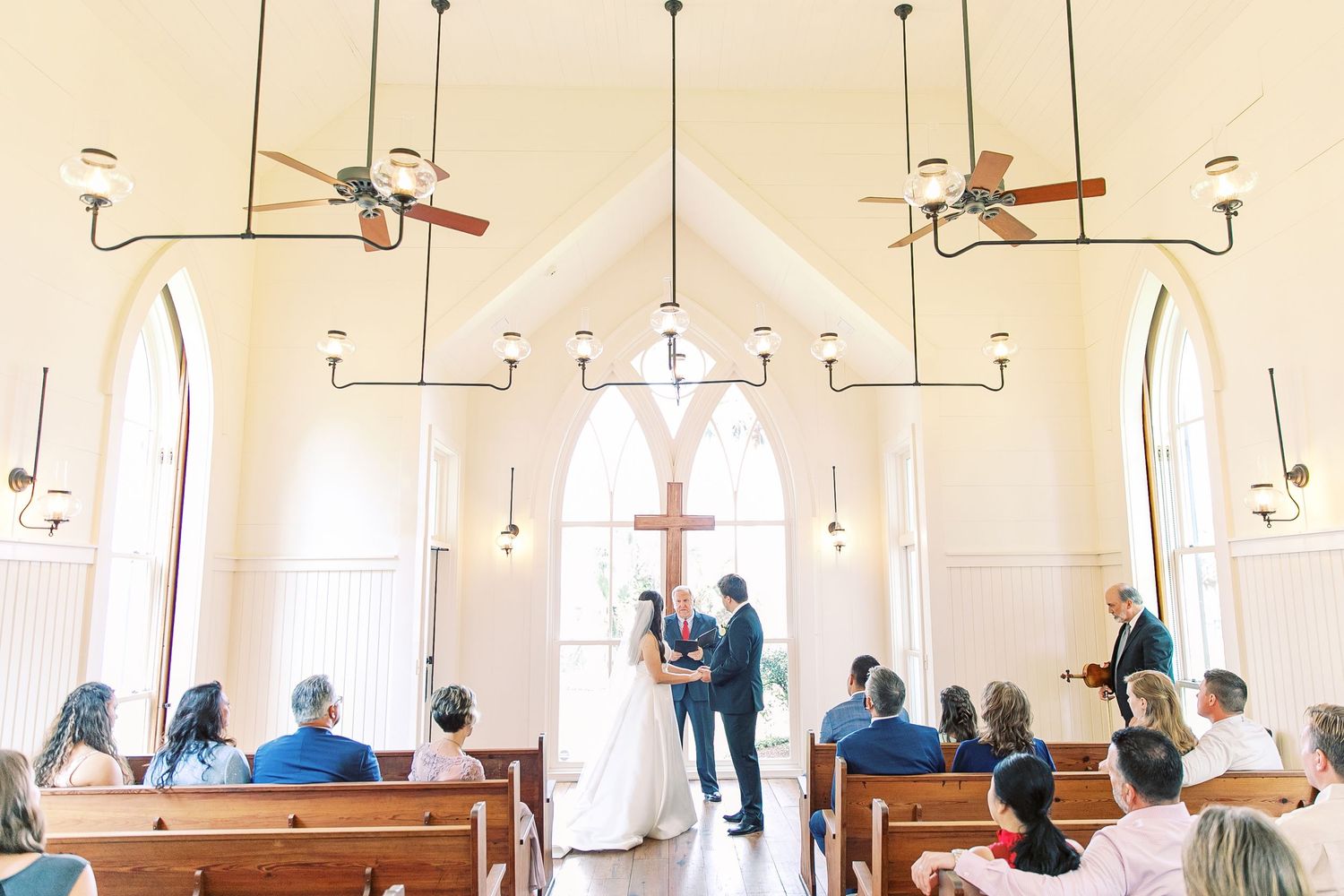 An intimate wedding ceremony takes place in a small white chapel with high ceilings and rustic wooden pews in palmetto bluff.