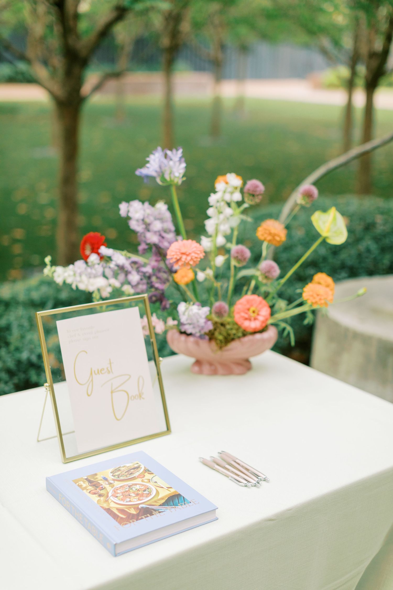 Wedding guestbook table with floral centerpiece and gold-framed sign on white tablecloth.