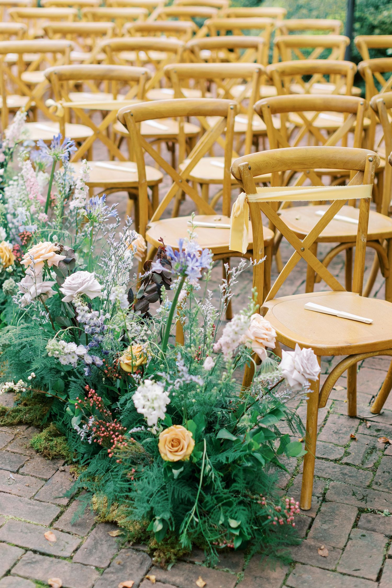 Rustic wooden chairs lined up for outdoor wedding ceremony with purple and peach floral aisle arrangements.