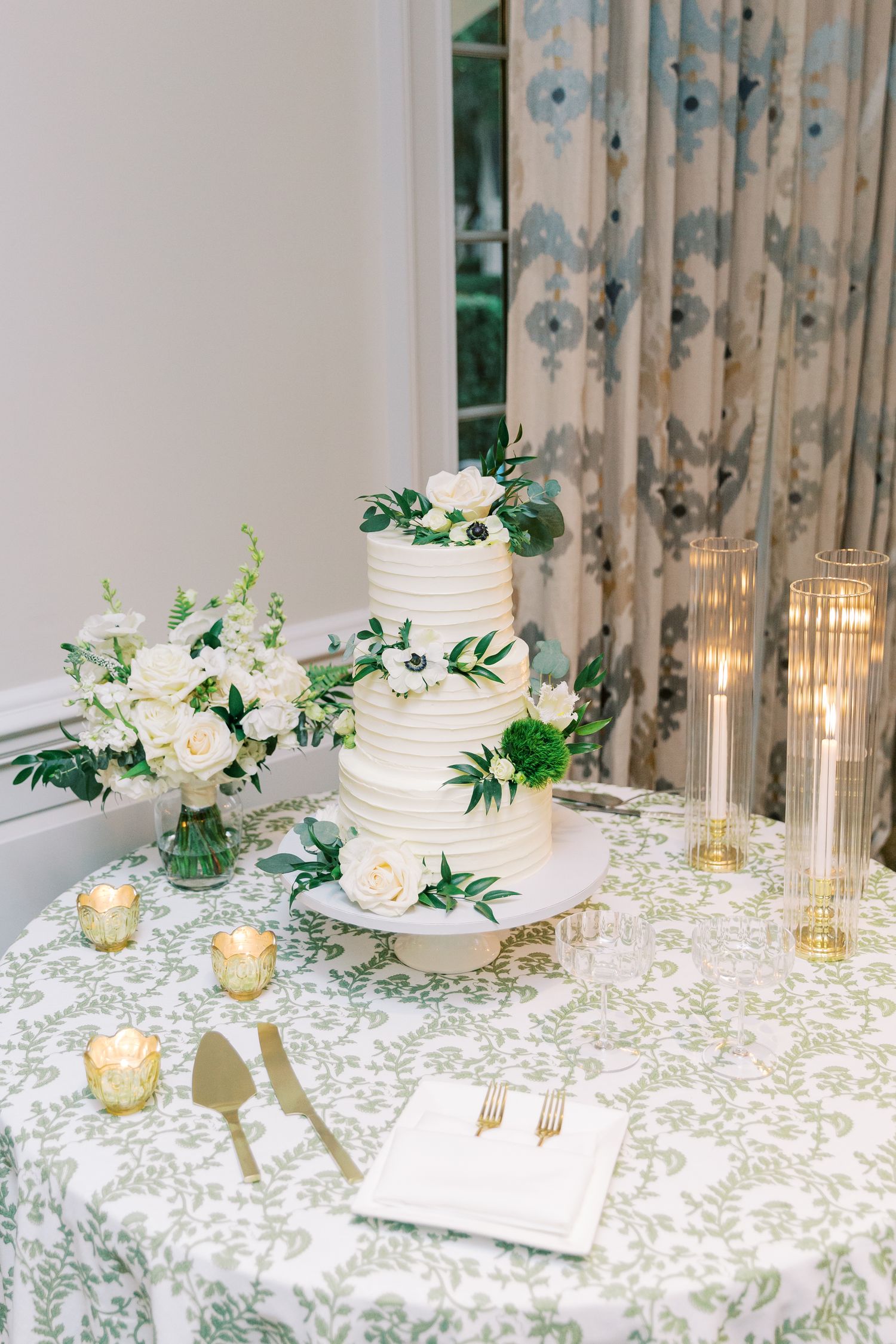 Elegant wedding table setting with white cake, floral arrangements, candles and patterned tablecloth in soft neutral tones.