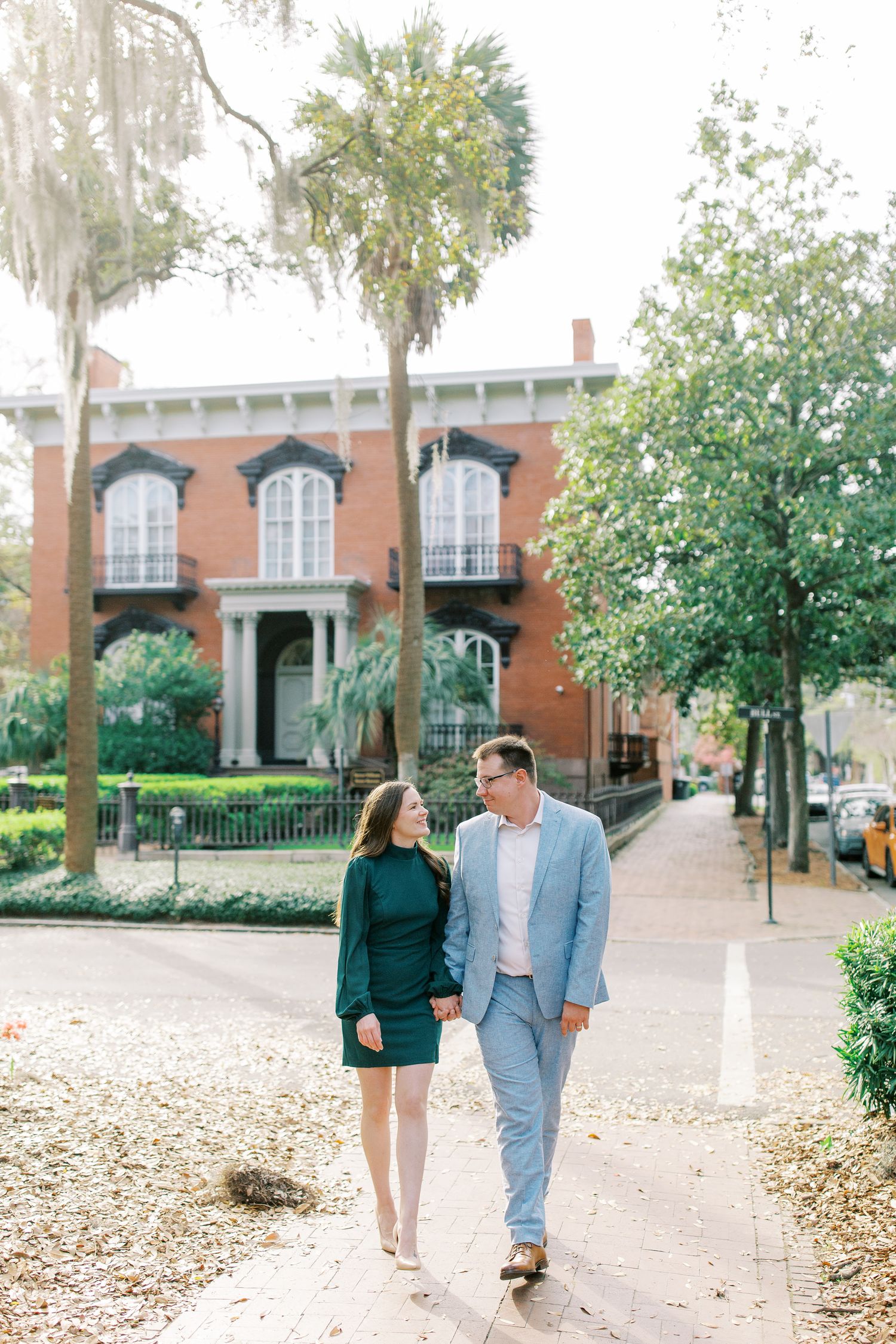 A couple strolling along a tree-lined path in front of a historic brick mansion with tall windows.
