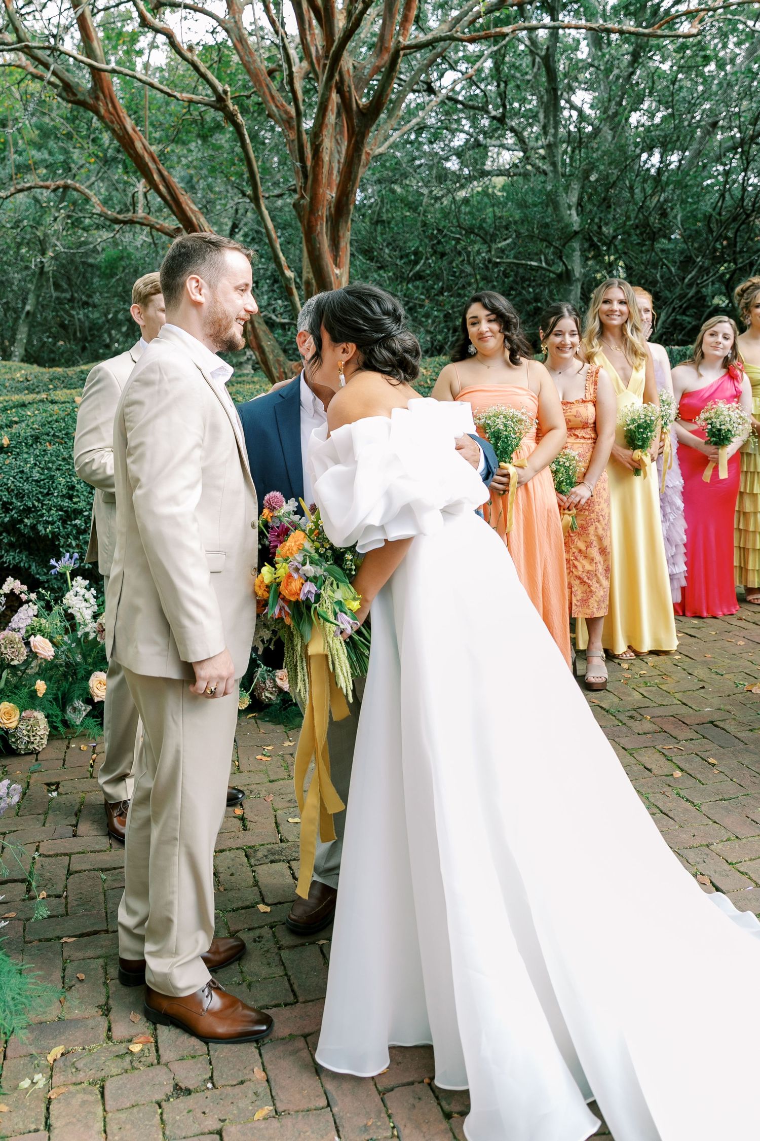 Wedding party in yellow and coral dresses gather for group photos on brick patio.