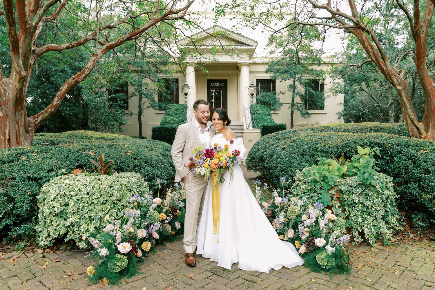 People in wedding attire gather for photos among lush green garden foliage and trees.