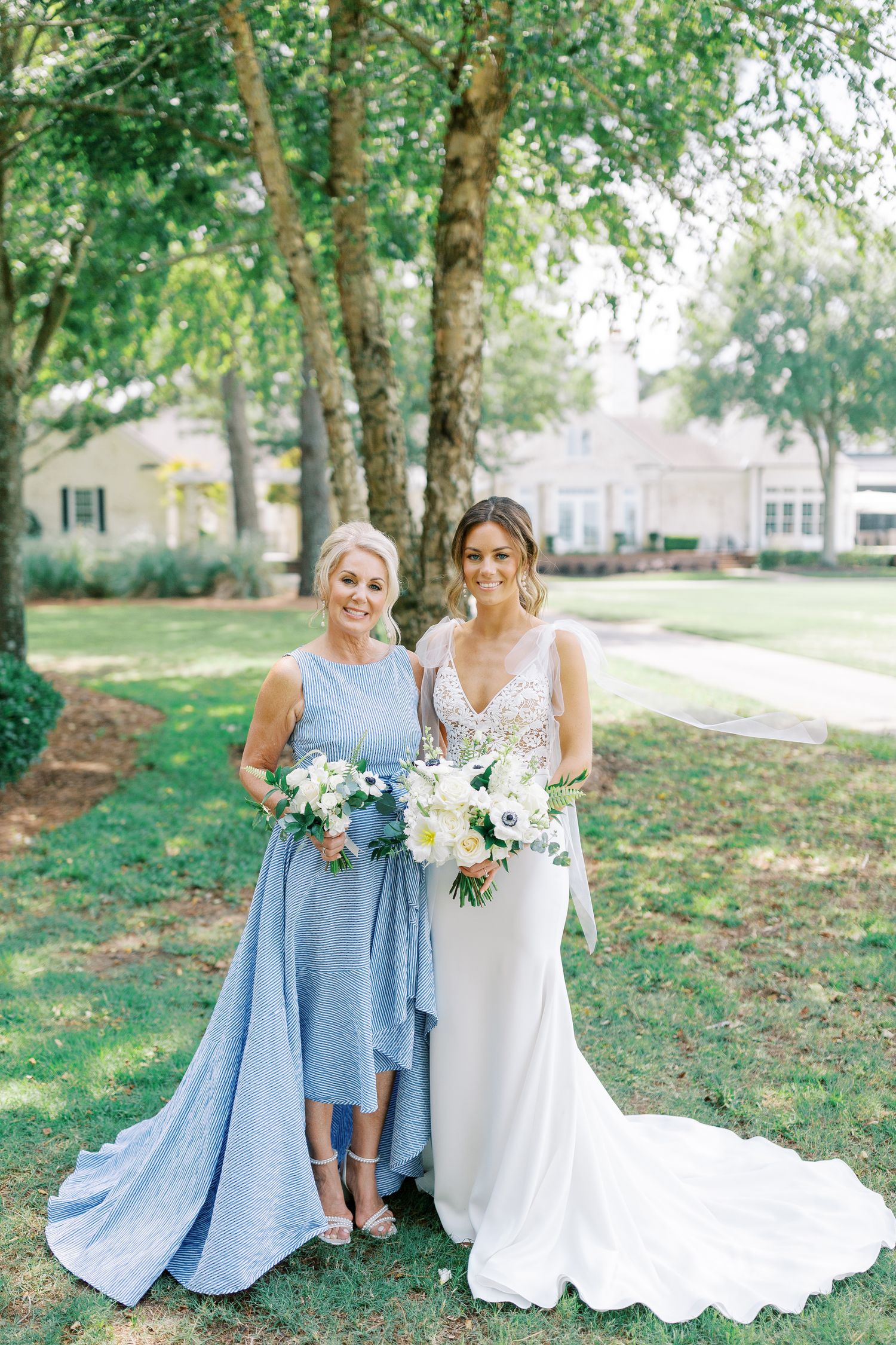 Bride and mom pose outdoors in elegant dresses, one in powder blue and one in white, holding floral bouquets on a lawn.