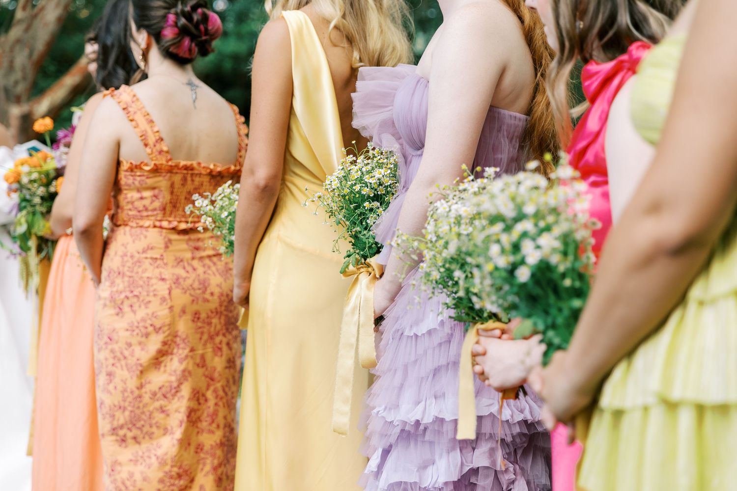 Row of bridesmaids in pastel peach, yellow and lavender dresses holding small floral bouquets at an outdoor wedding.