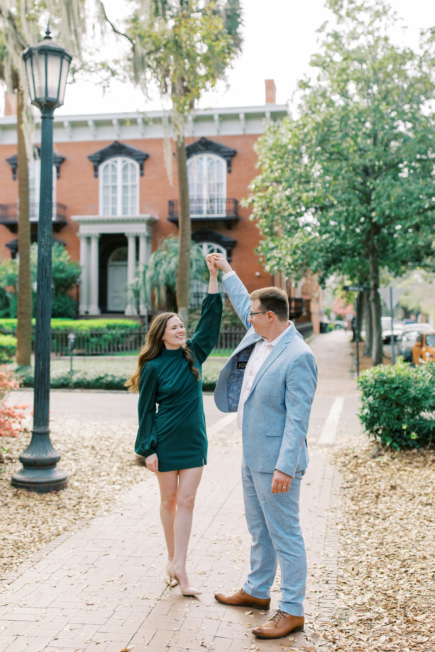 A couple dances playfully on a tree-lined brick pathway near historic buildings.
