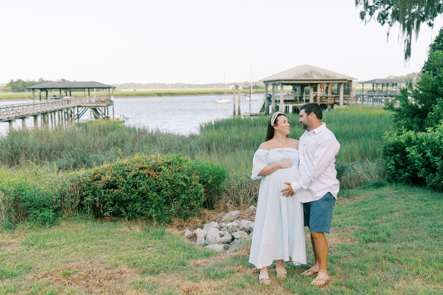 A couple embraces on a grassy lawn near the isle of hope waterfront with a wooden dock surrounded by coastal marshland.