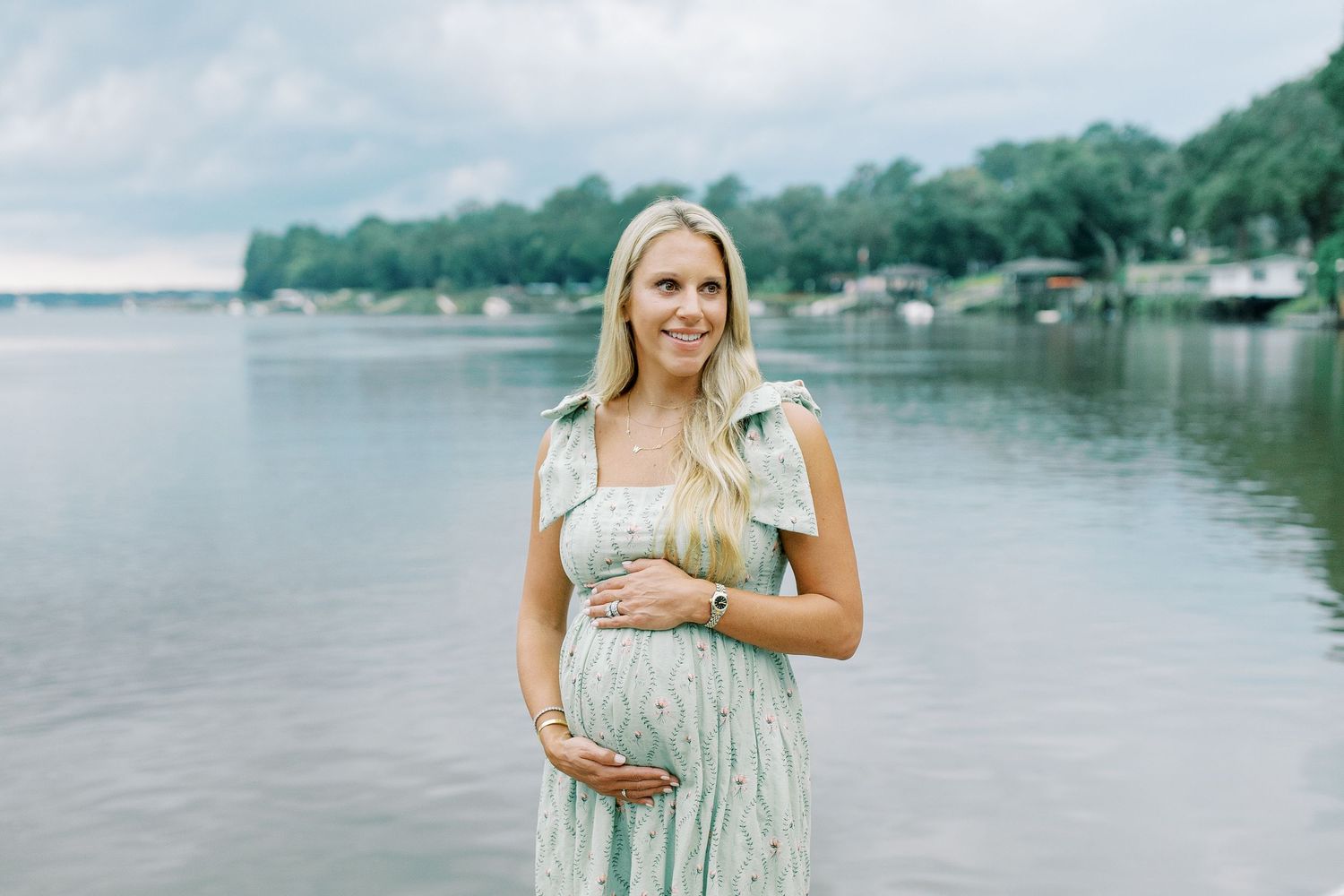 Expectant mother in light floral dress stands by peaceful lakeside during maternity photoshoot.