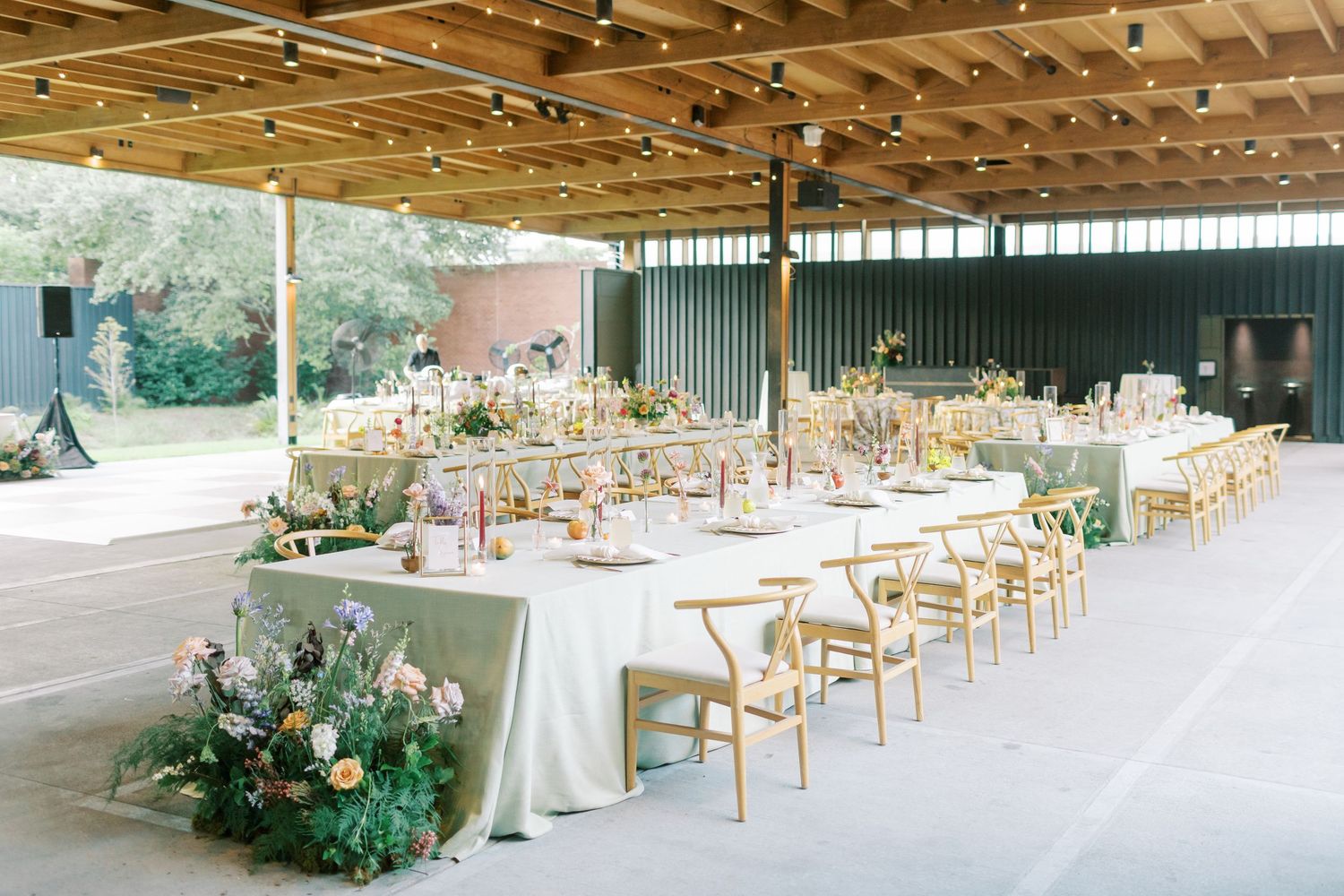 Elegant outdoor wedding reception setup with long tables, white linens, and natural wooden chairs under a covered pavilion.