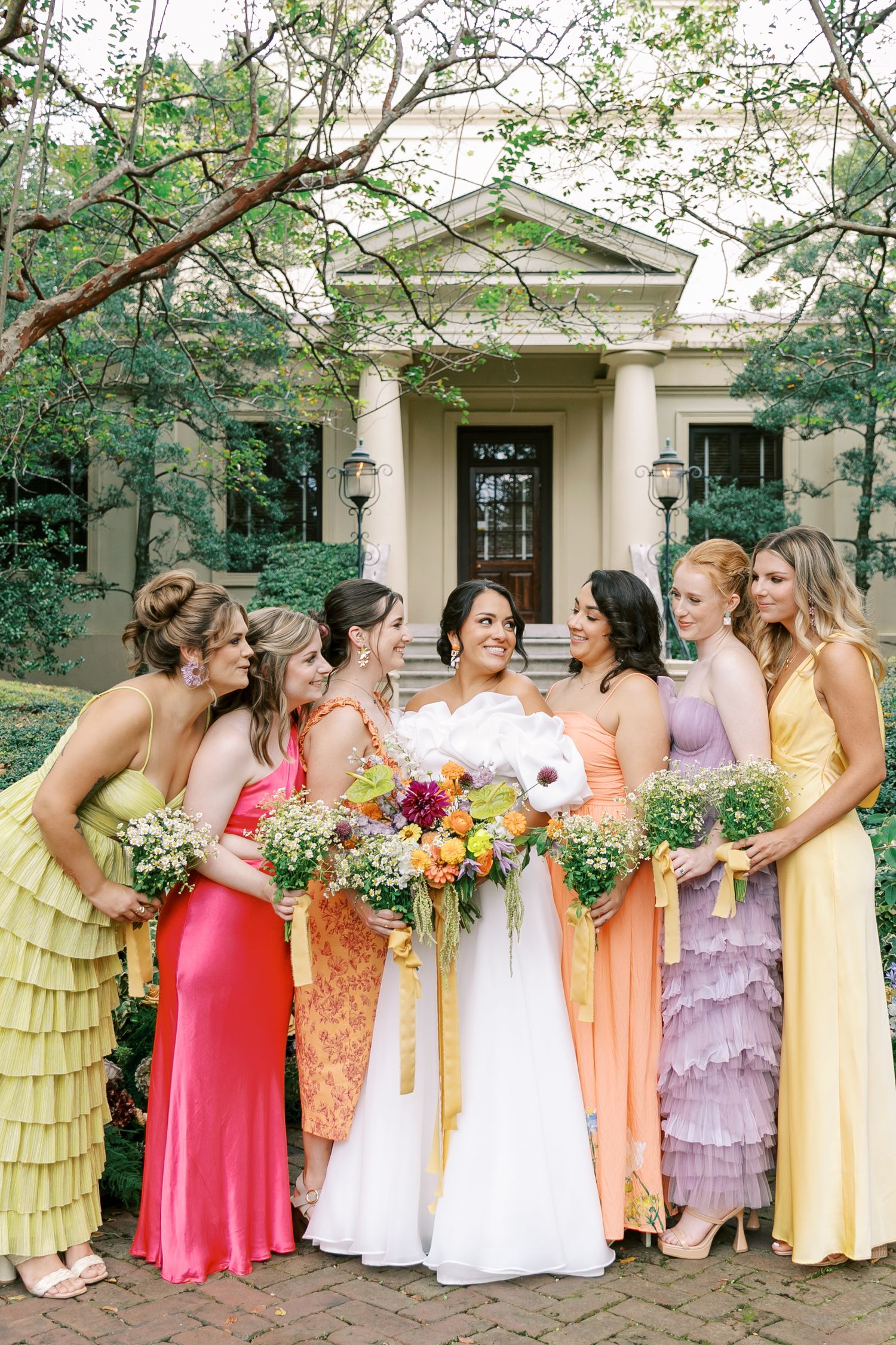 Bridesmaids wearing colorful pastel dresses line up in front of a classic building entrance.