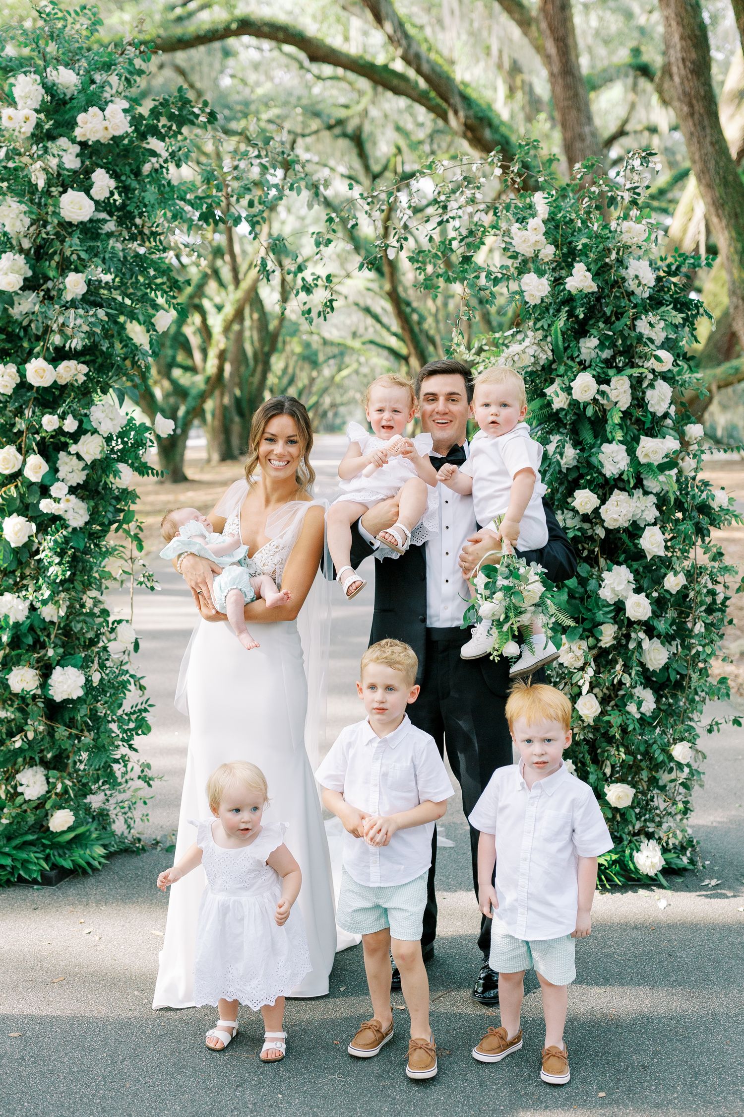 Elegant floral arch frames a family in formal attire against a backdrop of oak trees.