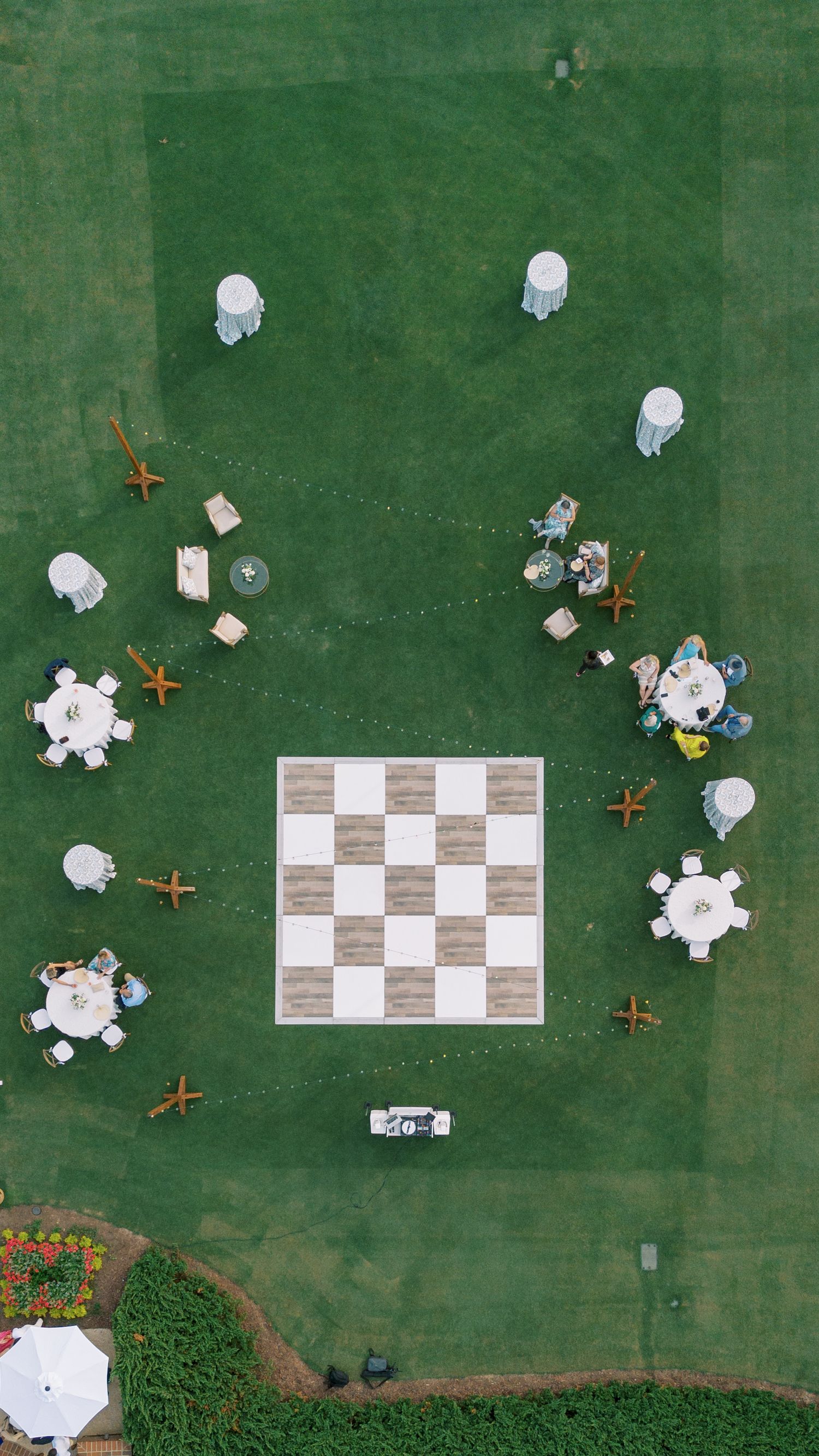 Aerial view of an outdoor event space with tables and a large decorative checkerboard pattern on green grass.