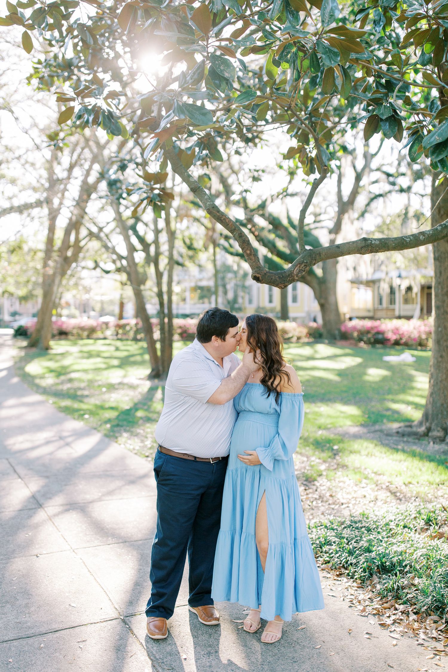 Couple sharing romantic moment outdoors in sunlit garden with person wearing flowing blue dress.