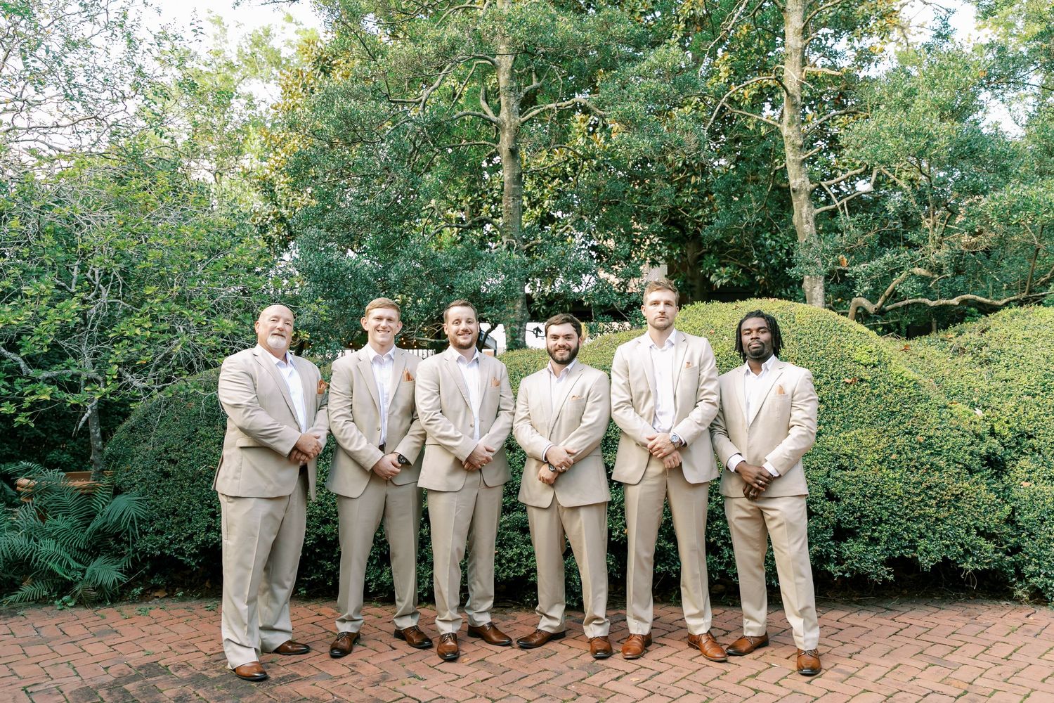 Groomsmen in matching beige suits pose together on garden brick pathway.