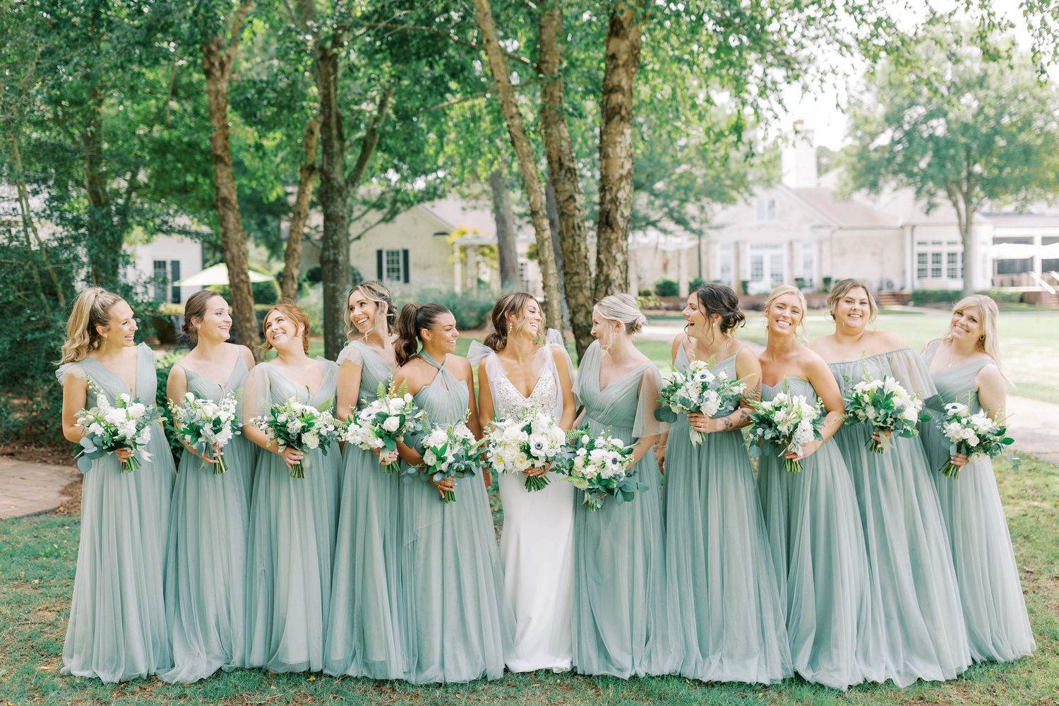 Bridal party in sage green chiffon dresses stands together on a lawn with white and green bouquets.