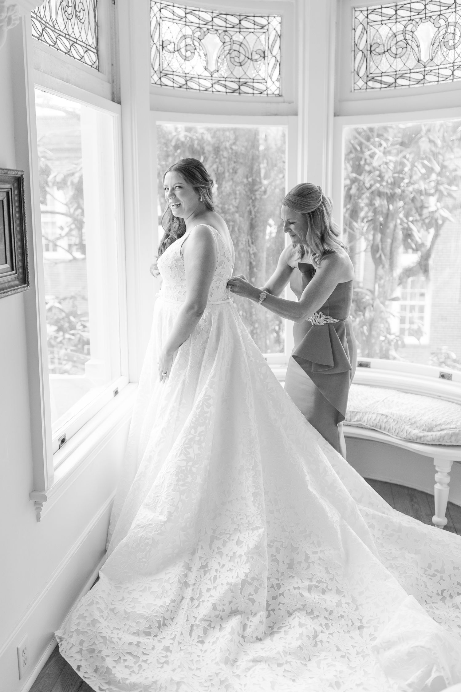 A black and white photograph shows a bride getting her wedding dress adjusted in an elegant bay window setting.