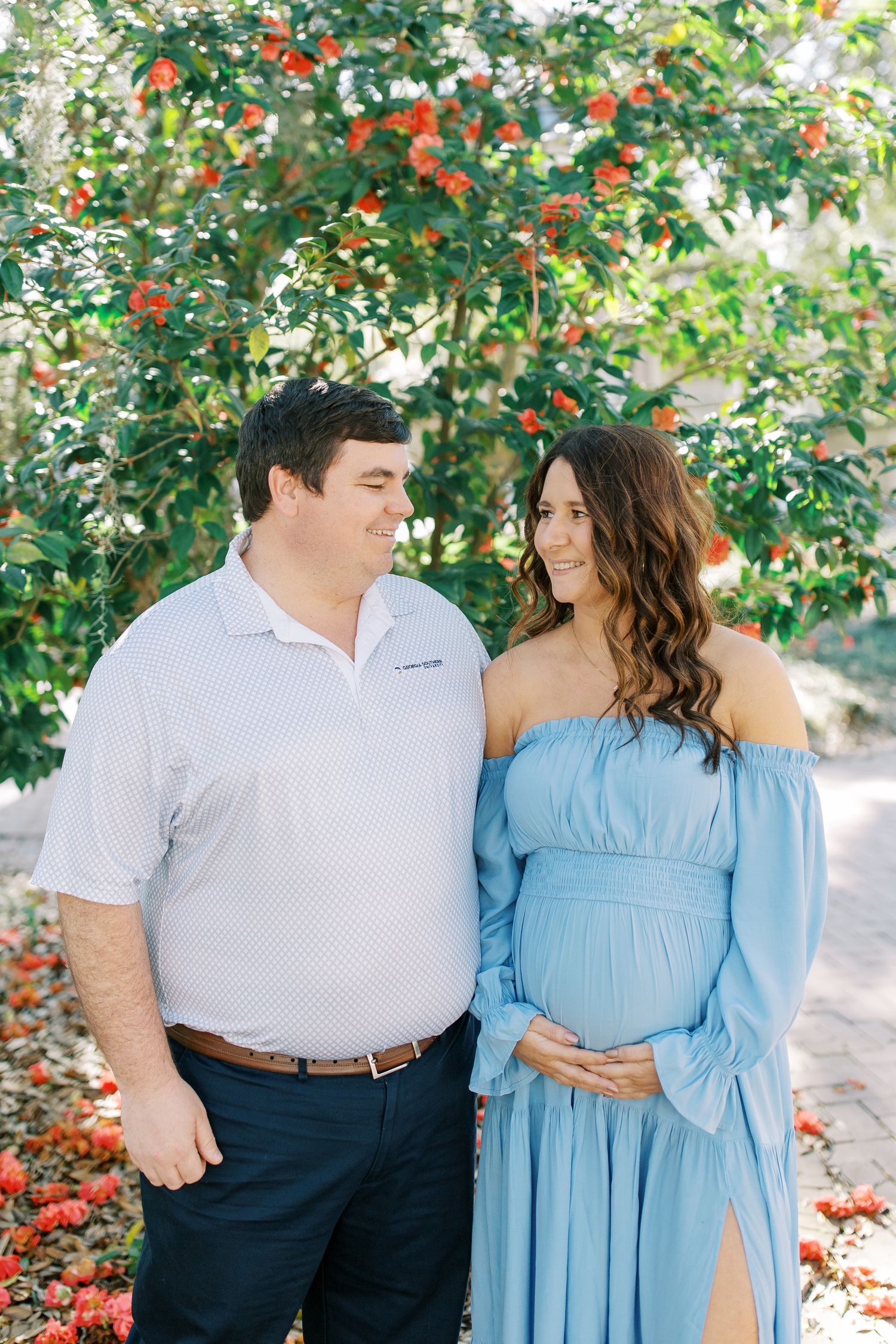 A couple stands under a flowering tree, with the woman wearing a light blue off-shoulder maternity dress.