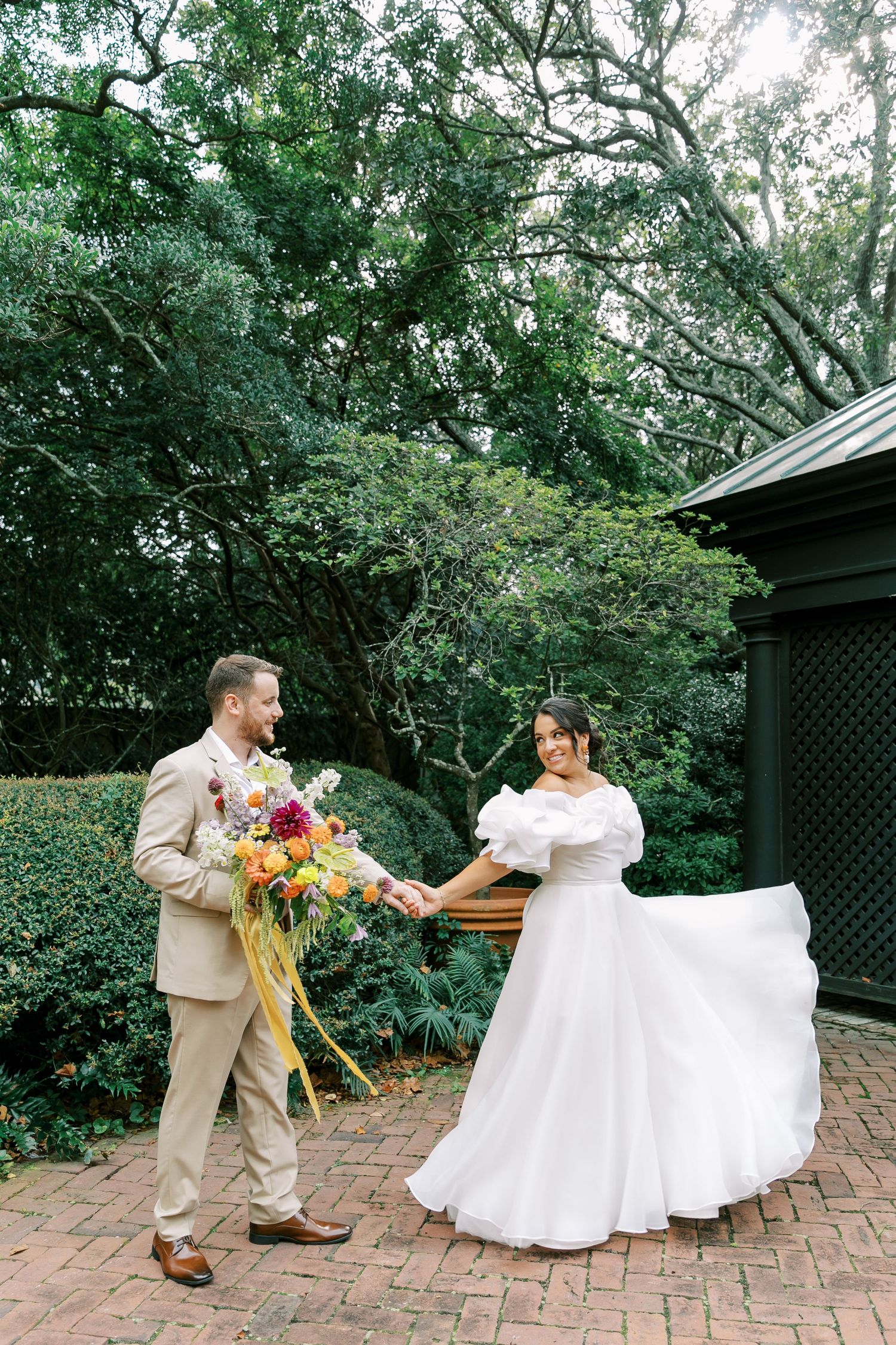 A couple sharing a romantic moment in a garden setting with the bride twirling in a white gown and groom holding flowers.