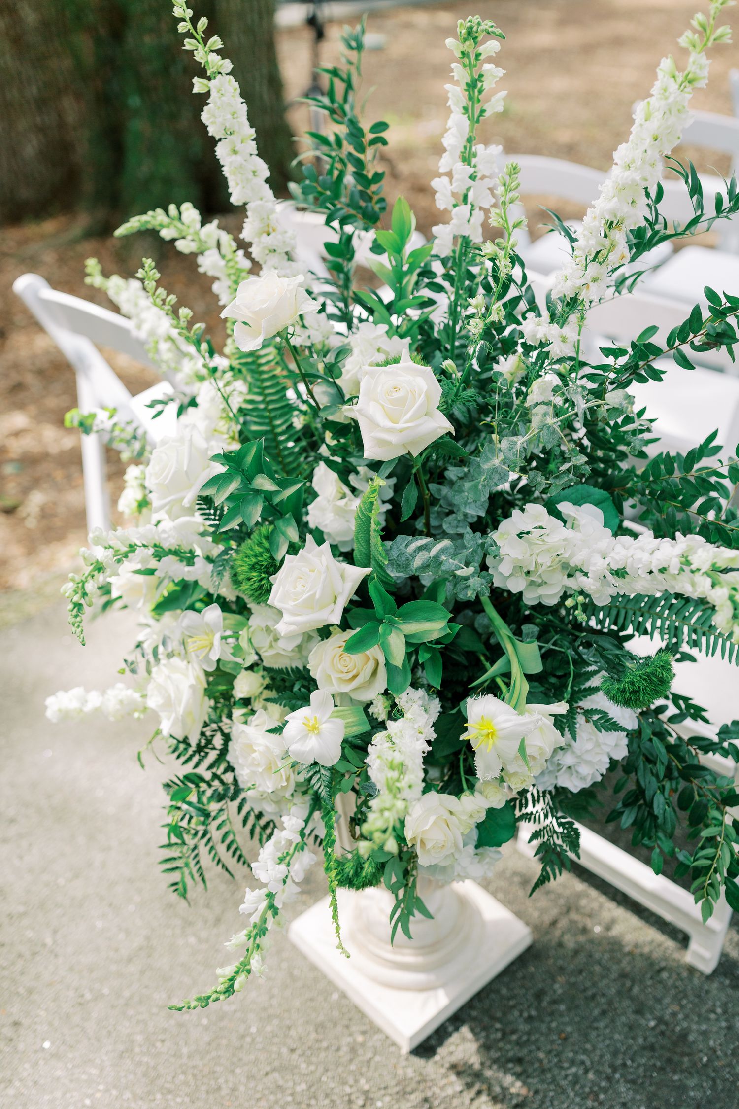 Elegant white floral arrangement featuring roses and delicate greenery in a classic white pedestal vase for a wedding ceremony.