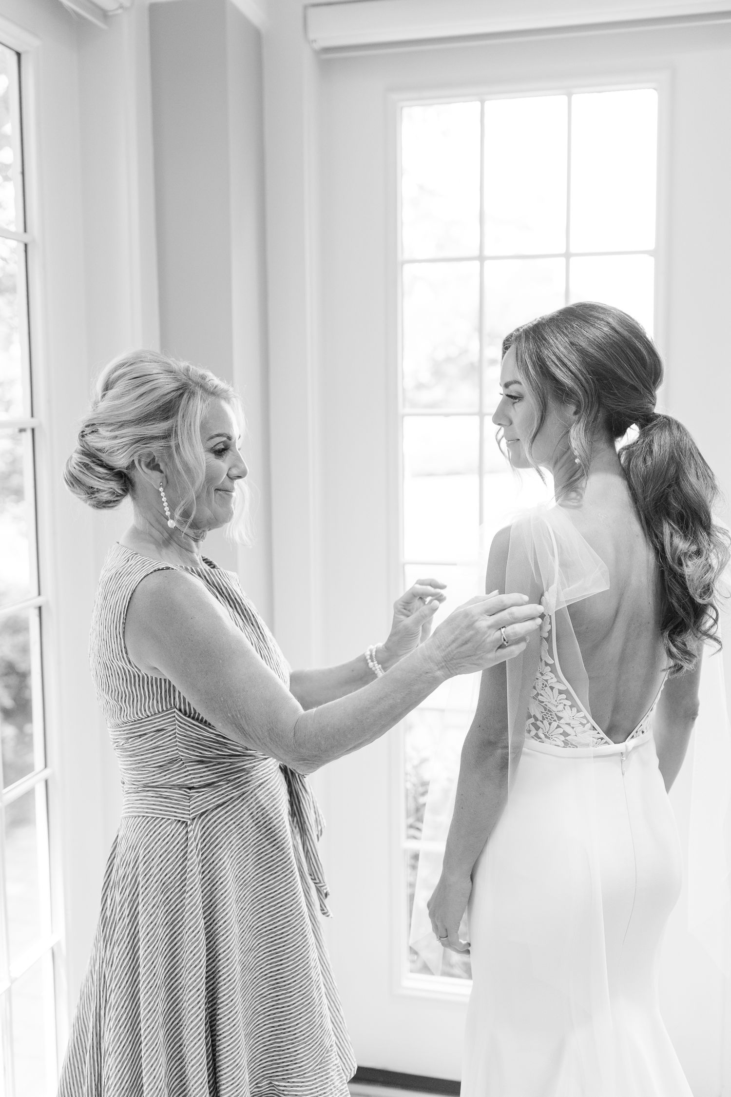 A tender moment captured in black and white as final adjustments are made to a stunning backless wedding gown by a window.