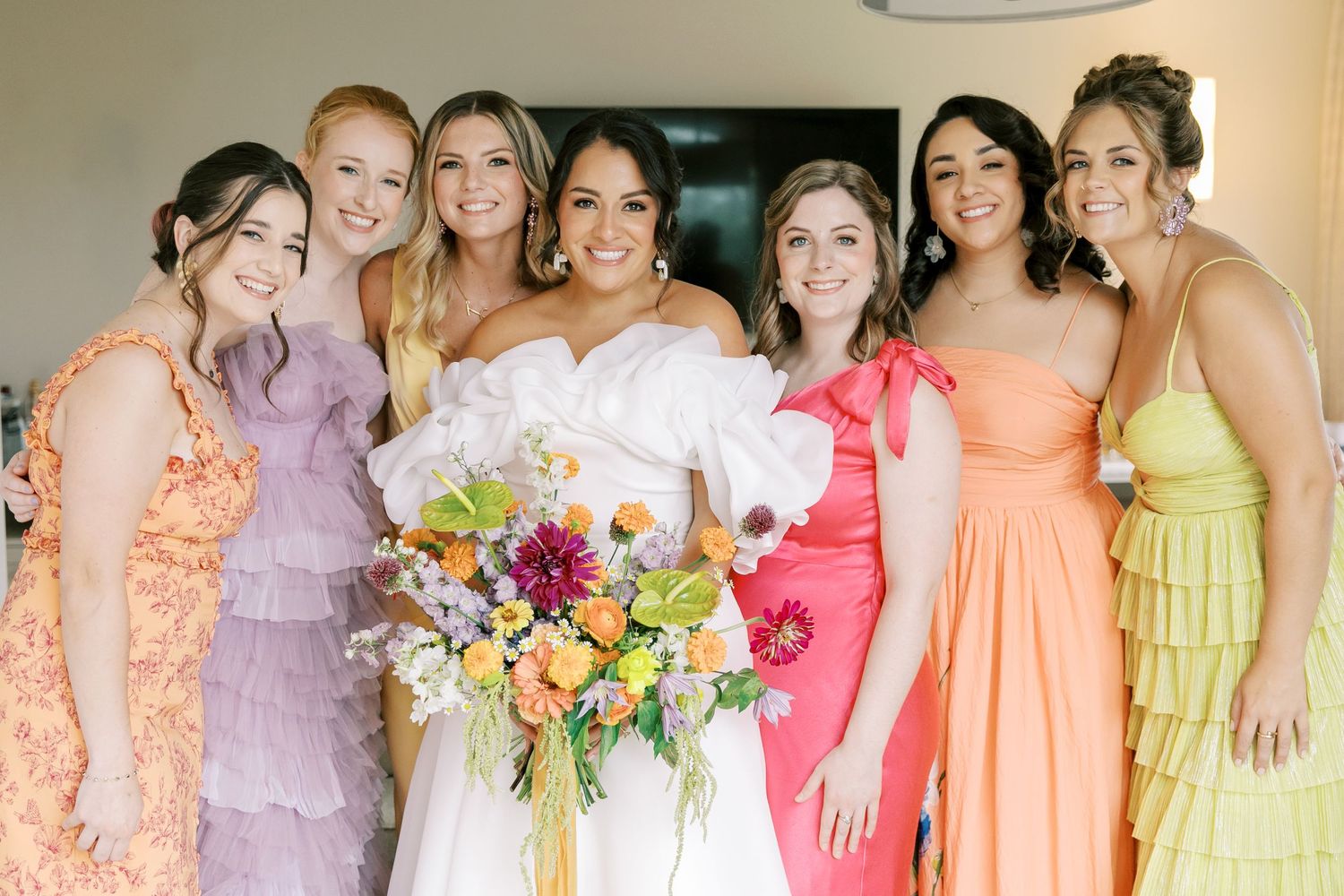 A bridal party wearing pastel dresses in lavender, coral, pink and yellow poses together with a colorful bouquet.