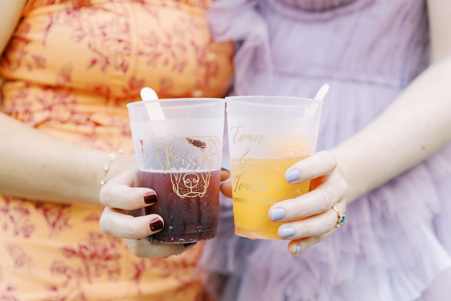 Two hands holding colorful beverages in clear plastic cups at a wedding celebration.