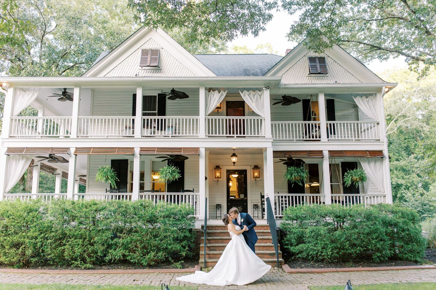 Two people pose in front of an elegant white plantation-style house with double-story wraparound porches and green landscaping.