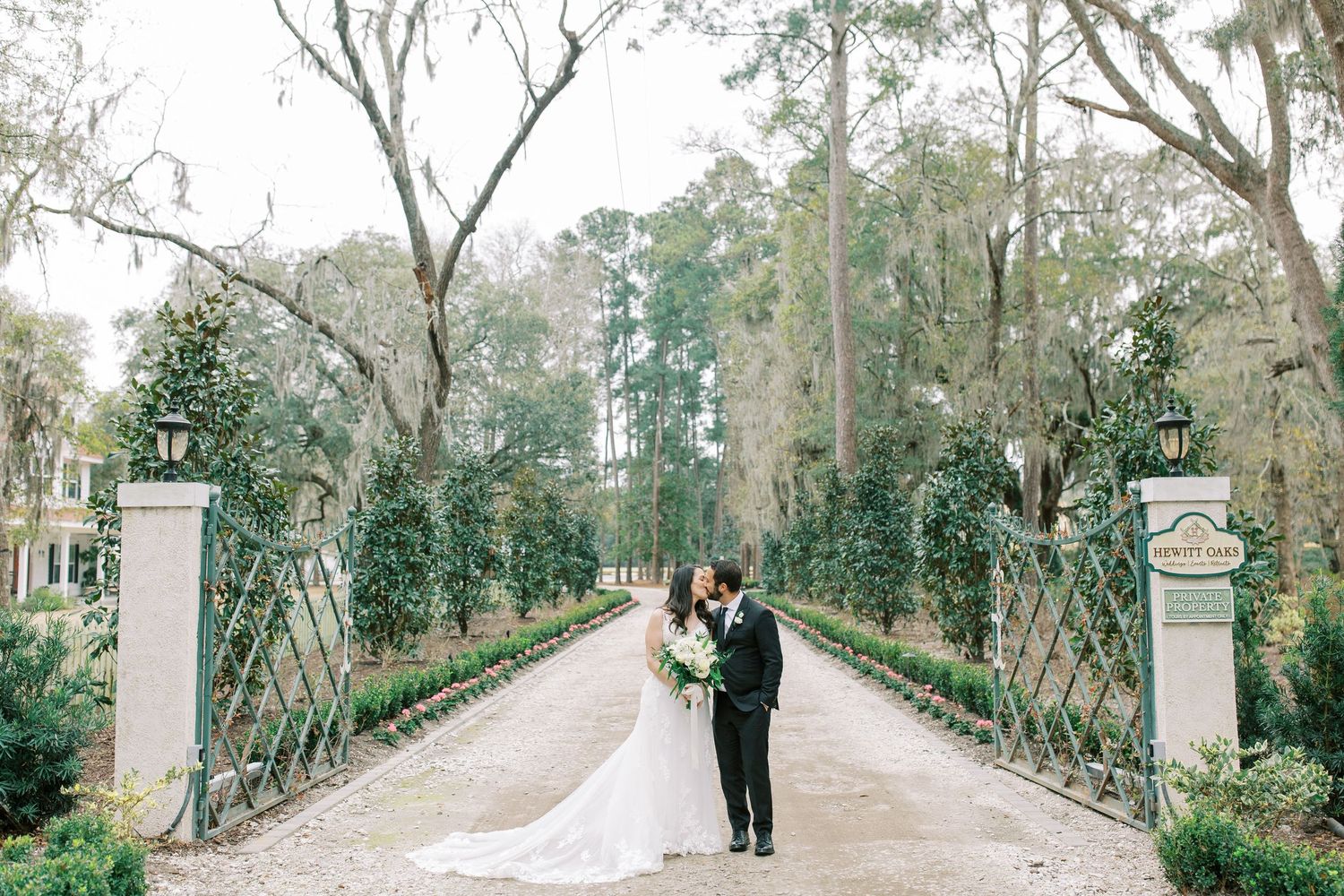 A couple enjoys a romantic moment on a tree-lined garden path between ornate stone pillars with lanterns at Hewitt Oaks in Bluffton, SC.