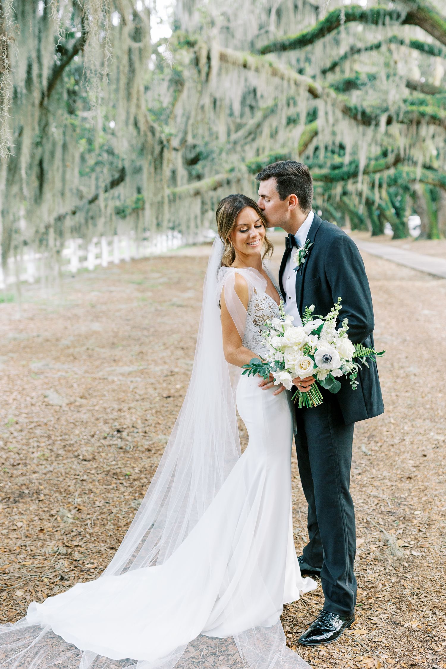 Wedding couple embraces under Spanish moss trees with bride in flowing white gown holding floral bouquet.