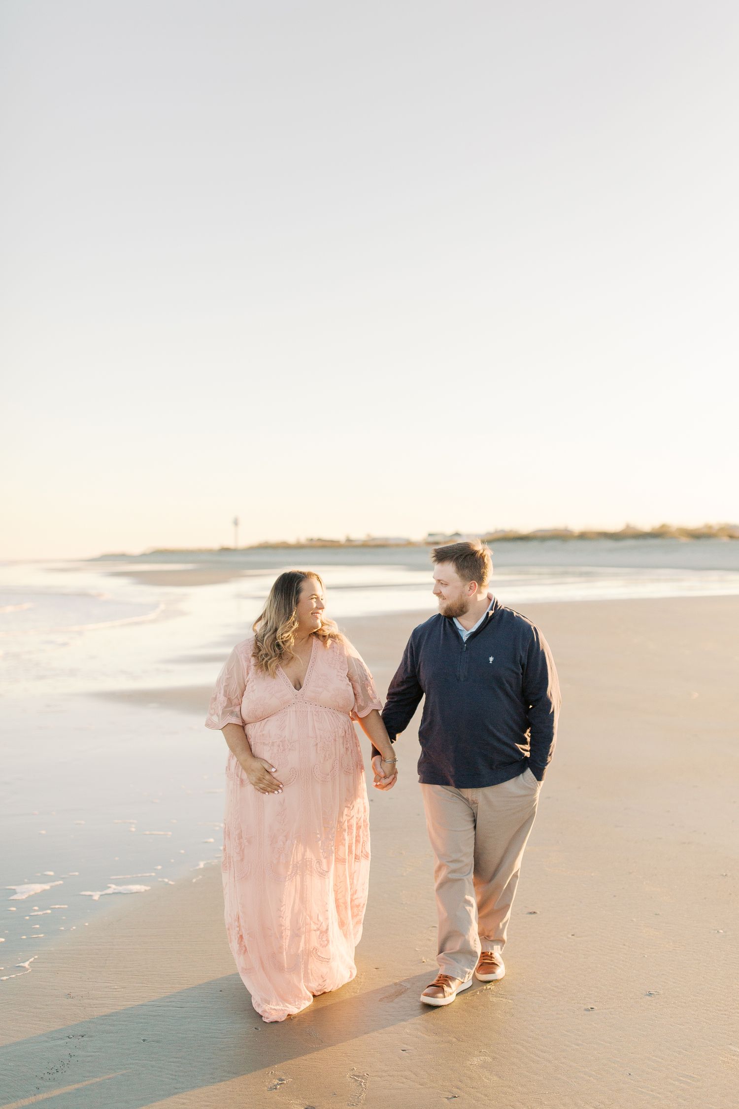 A couple walks hand in hand along a peaceful beach at sunset, with her in a flowing pink dress and him in navy blue.