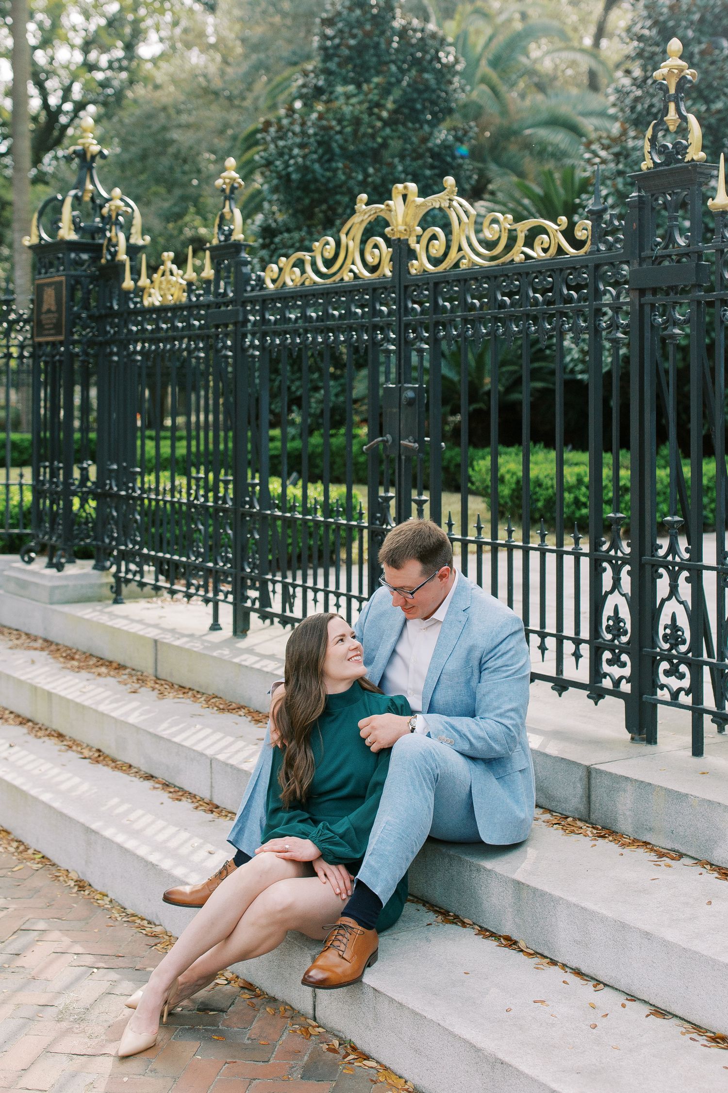 Romantic couple in green and blue sits together on white stone steps beside decorative black iron fence.