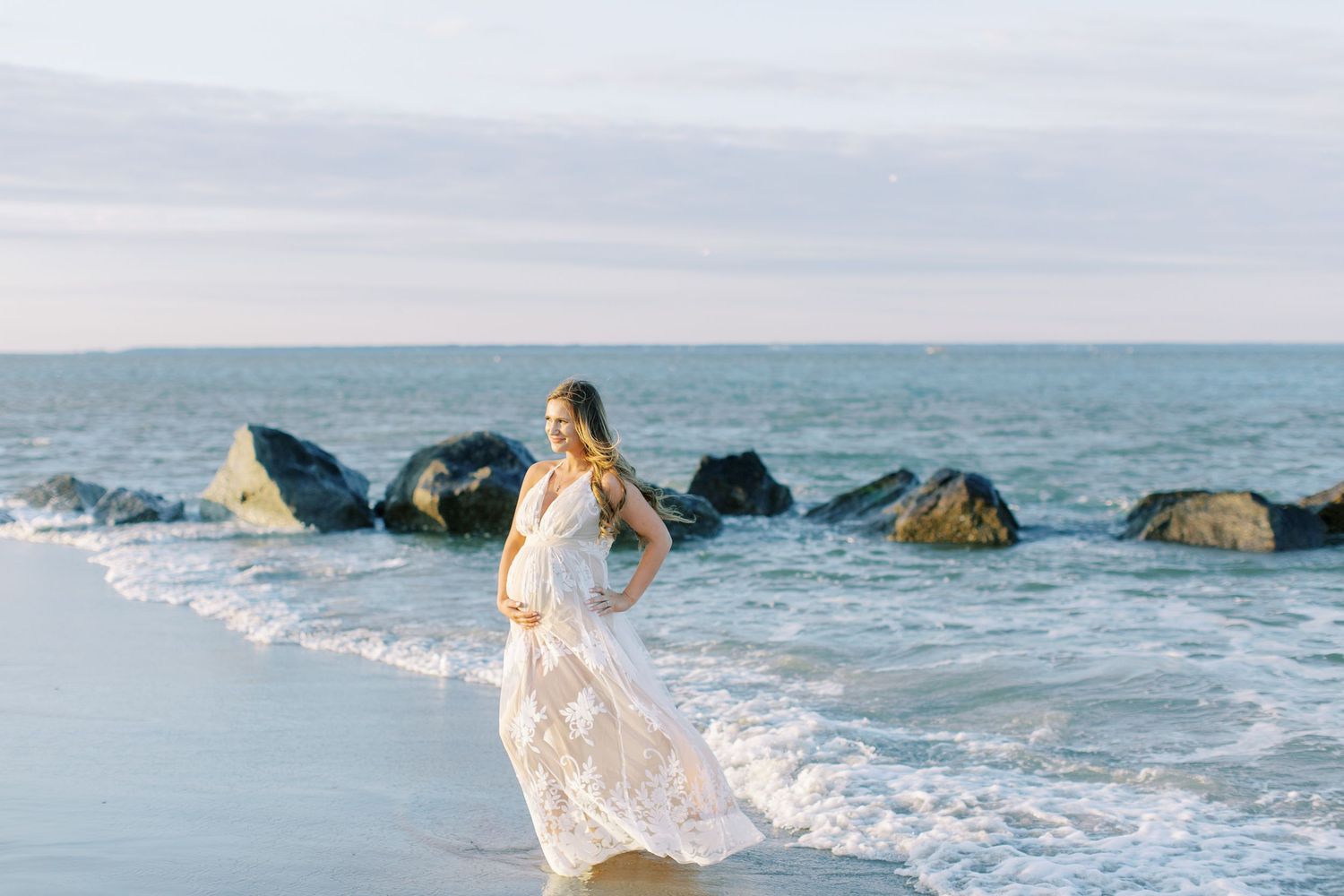 A flowing white lace dress catches the ocean breeze as waves crash against rocky shores on a cloudy beach day on tybee island.
