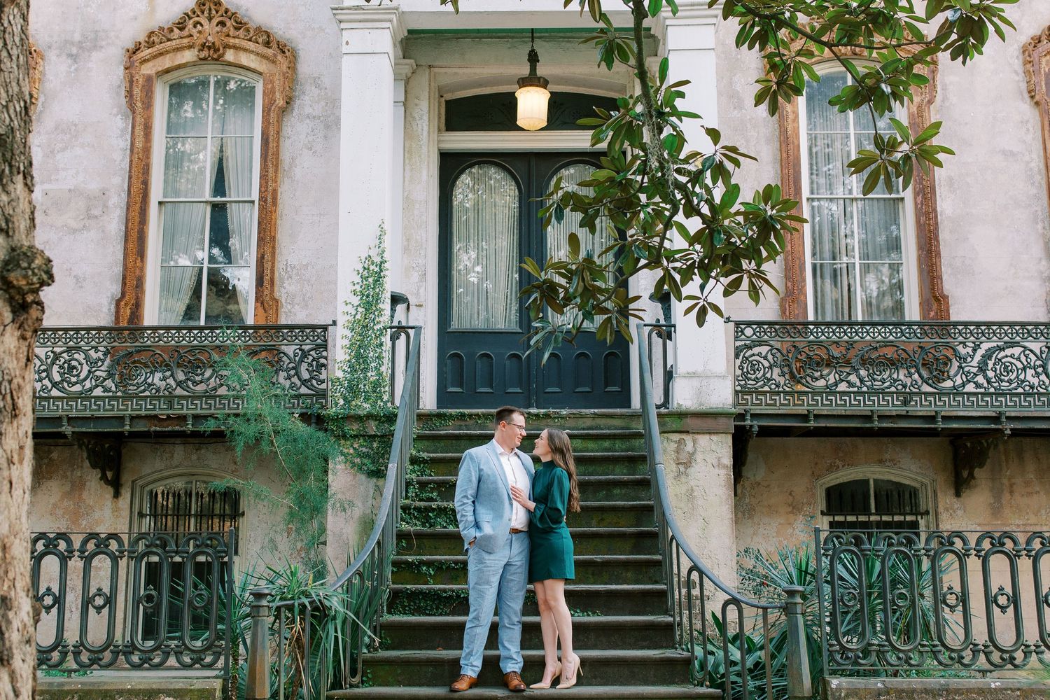 A romantic couple stands on ornate wrought iron steps leading to a historic Victorian-style house in Savannah, Georgia.