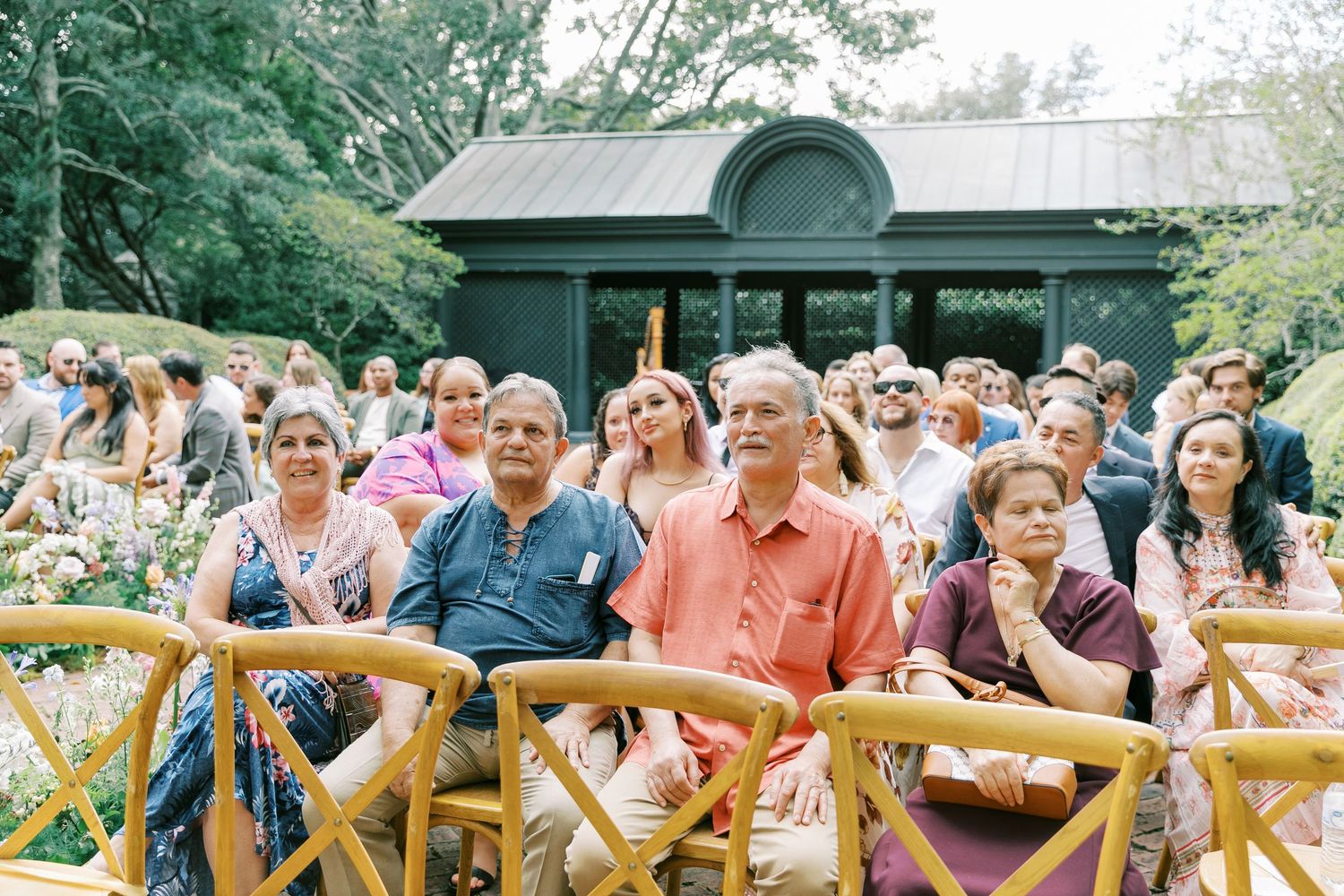 Wedding guests seated on wooden chairs watching ceremony in an outdoor garden gazebo setting.