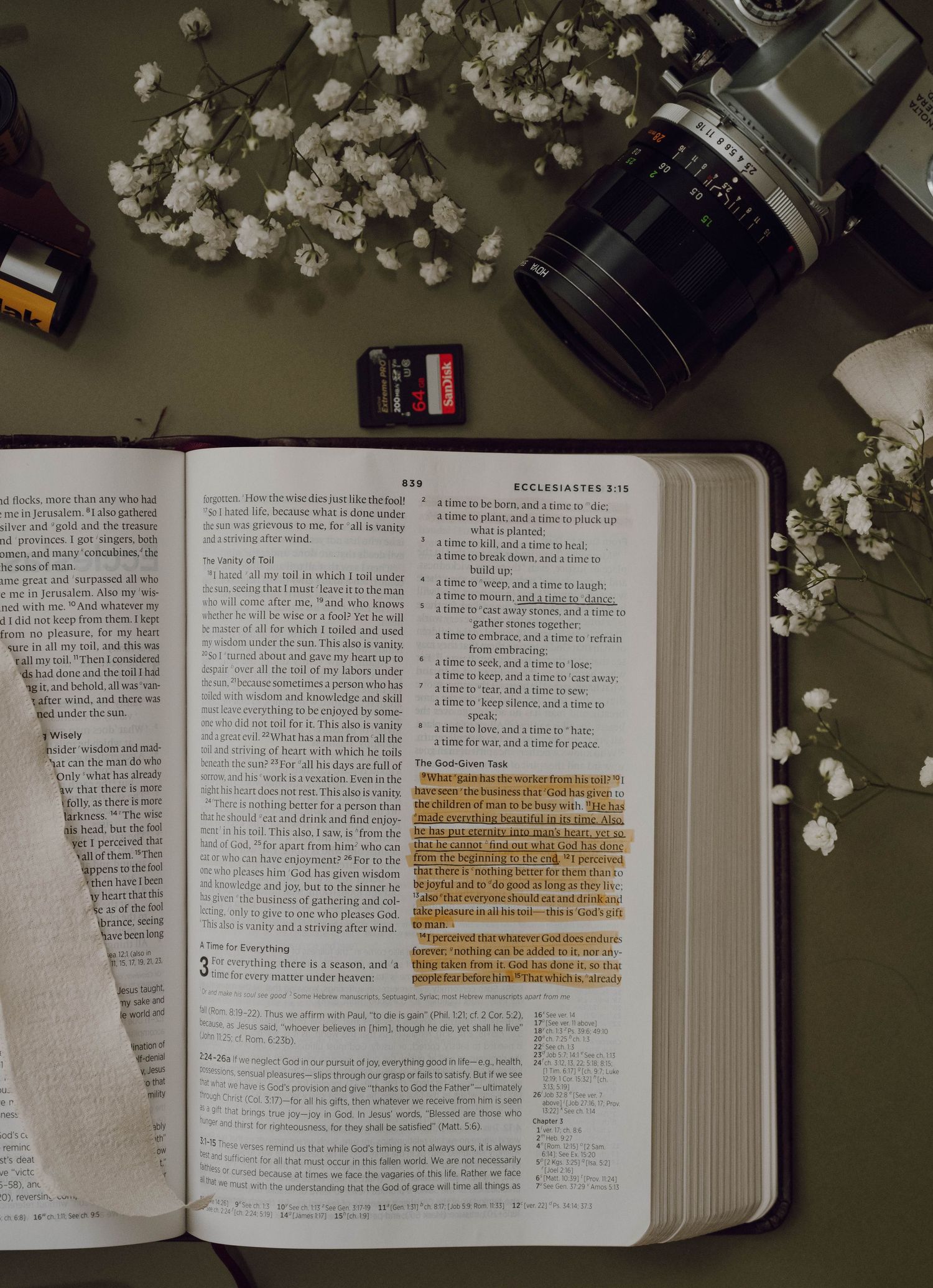 An open Bible with highlighted text surrounded by white baby's breath flowers and camera equipment on a green surface.