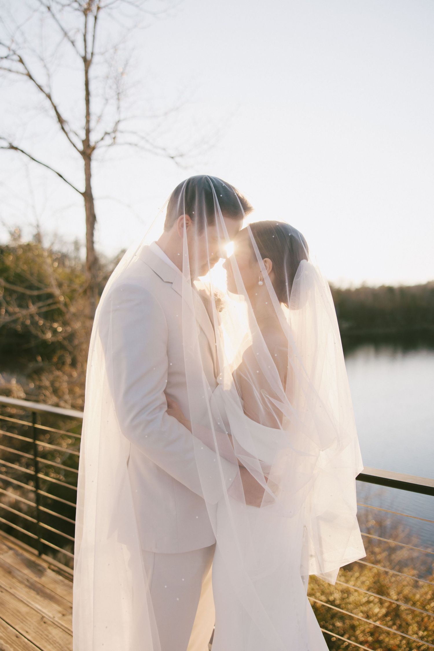 Newlywed couple sharing an intimate moment under a flowing veil on a lakeside deck at sunset.