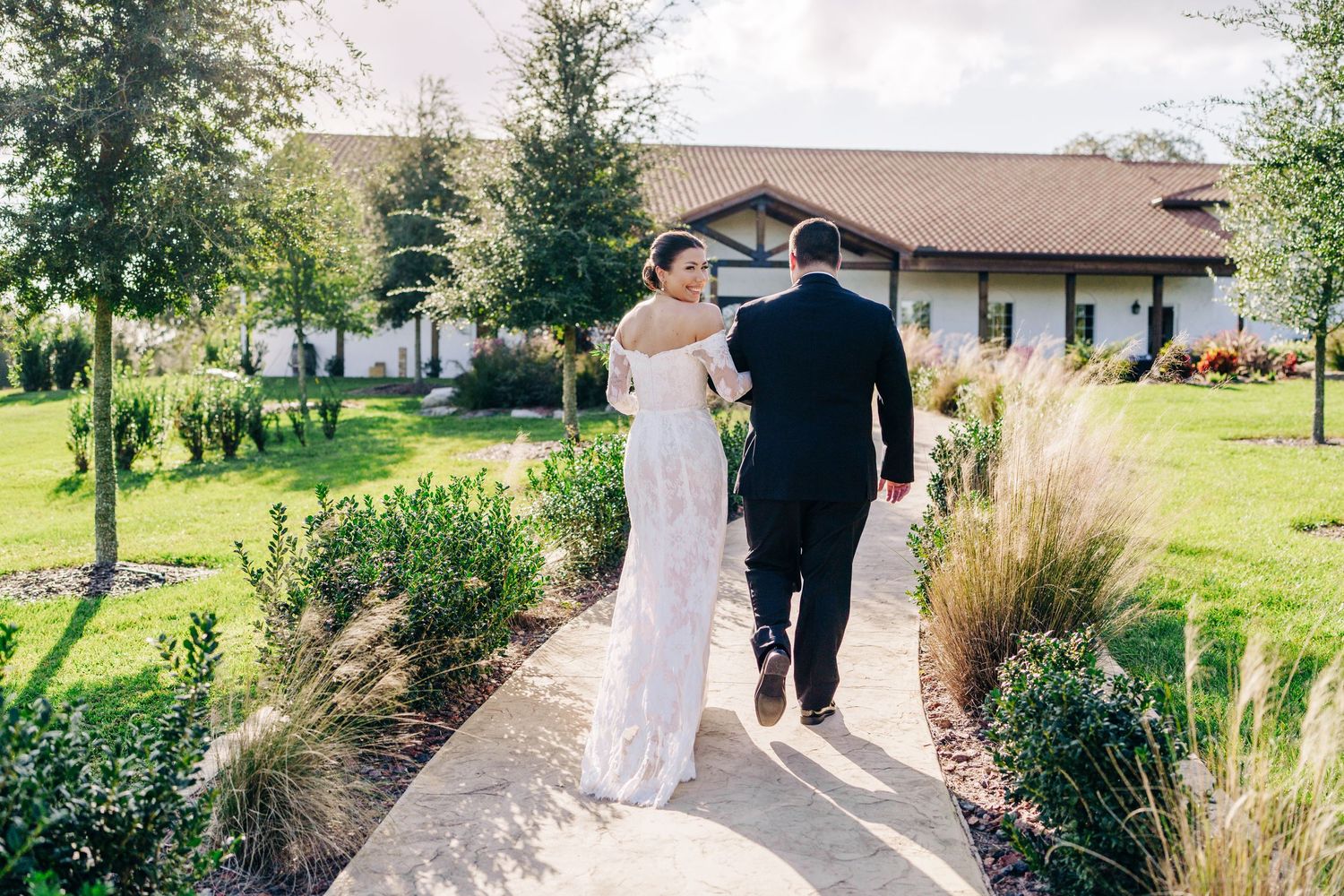 Couple walking down garden path at outdoor wedding venue with rustic ranch house and ornamental grasses.