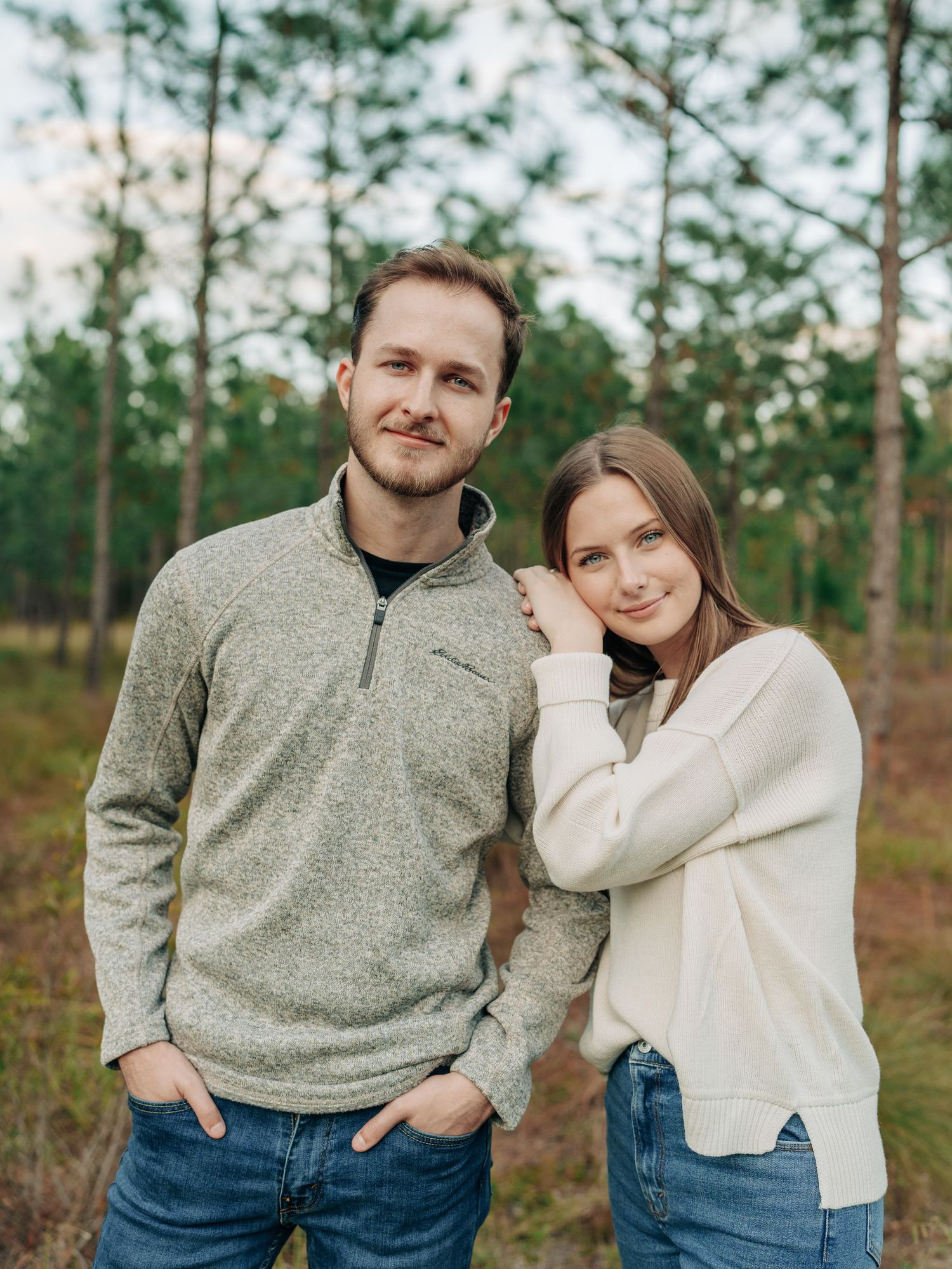 A couple in cozy sweaters poses together in a forest setting with pine trees in the background.