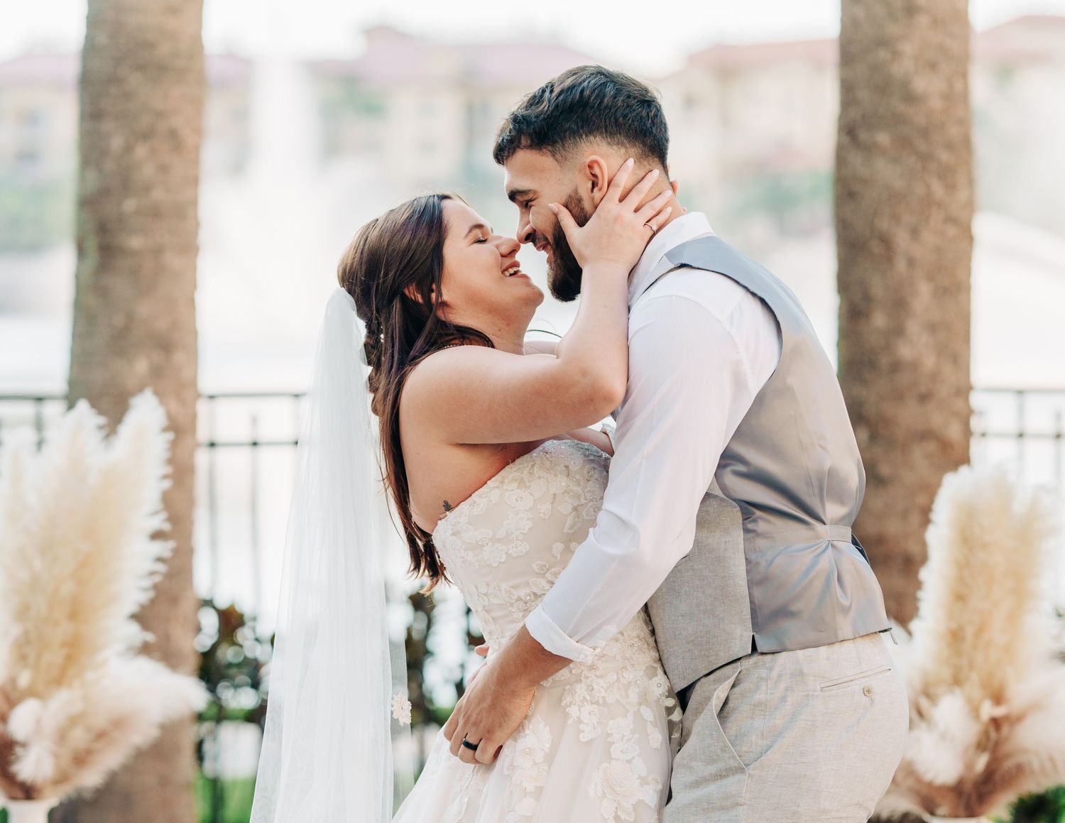 A romantic wedding couple embraces outdoors among palm trees and pampas grass in soft natural lighting.